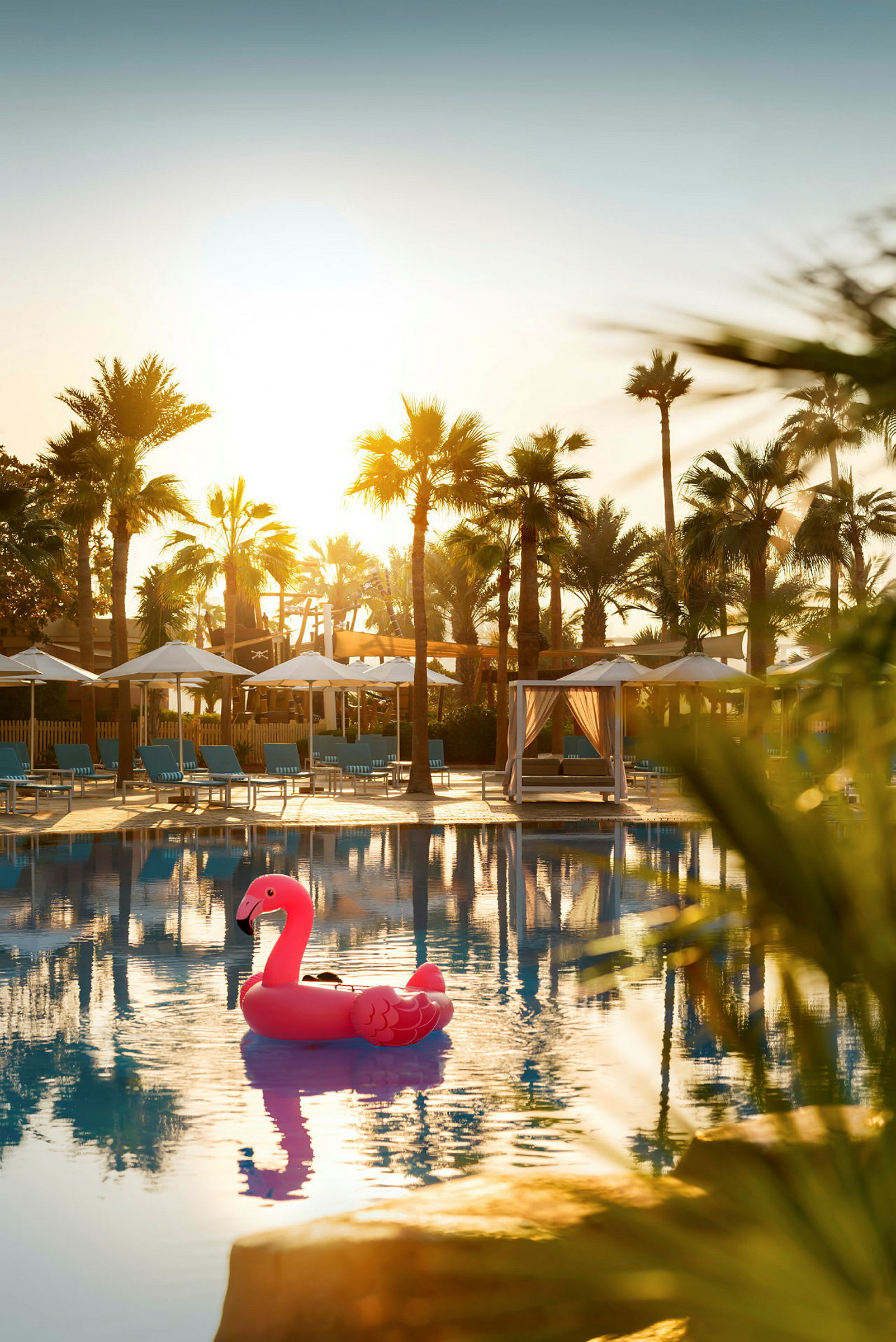 Rosa oppustelig flamingo i hotelpool med palmer og liggestole i baggrunden. (AI-genereret tekst)