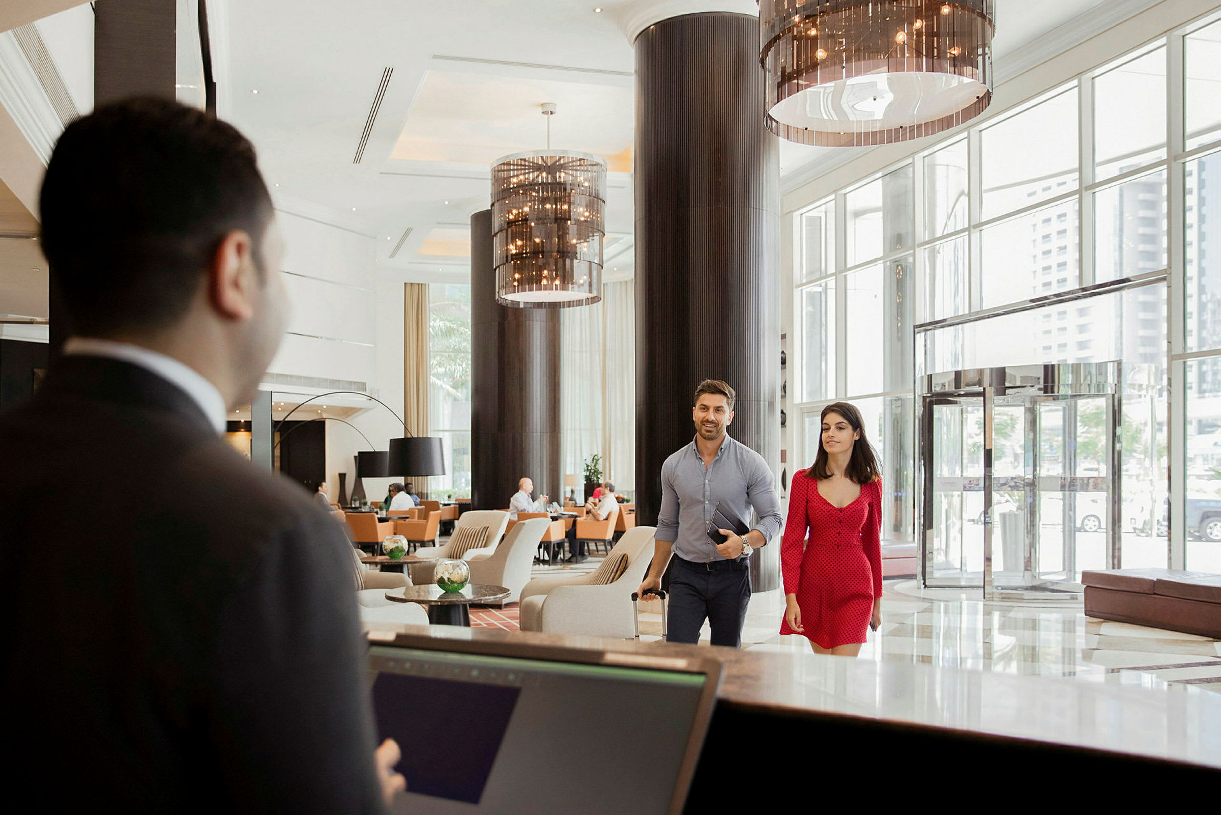 Interiør af en hotellobby med en reception i forgrunden og to gæster, der går mod receptionen. (AI-genereret tekst)
