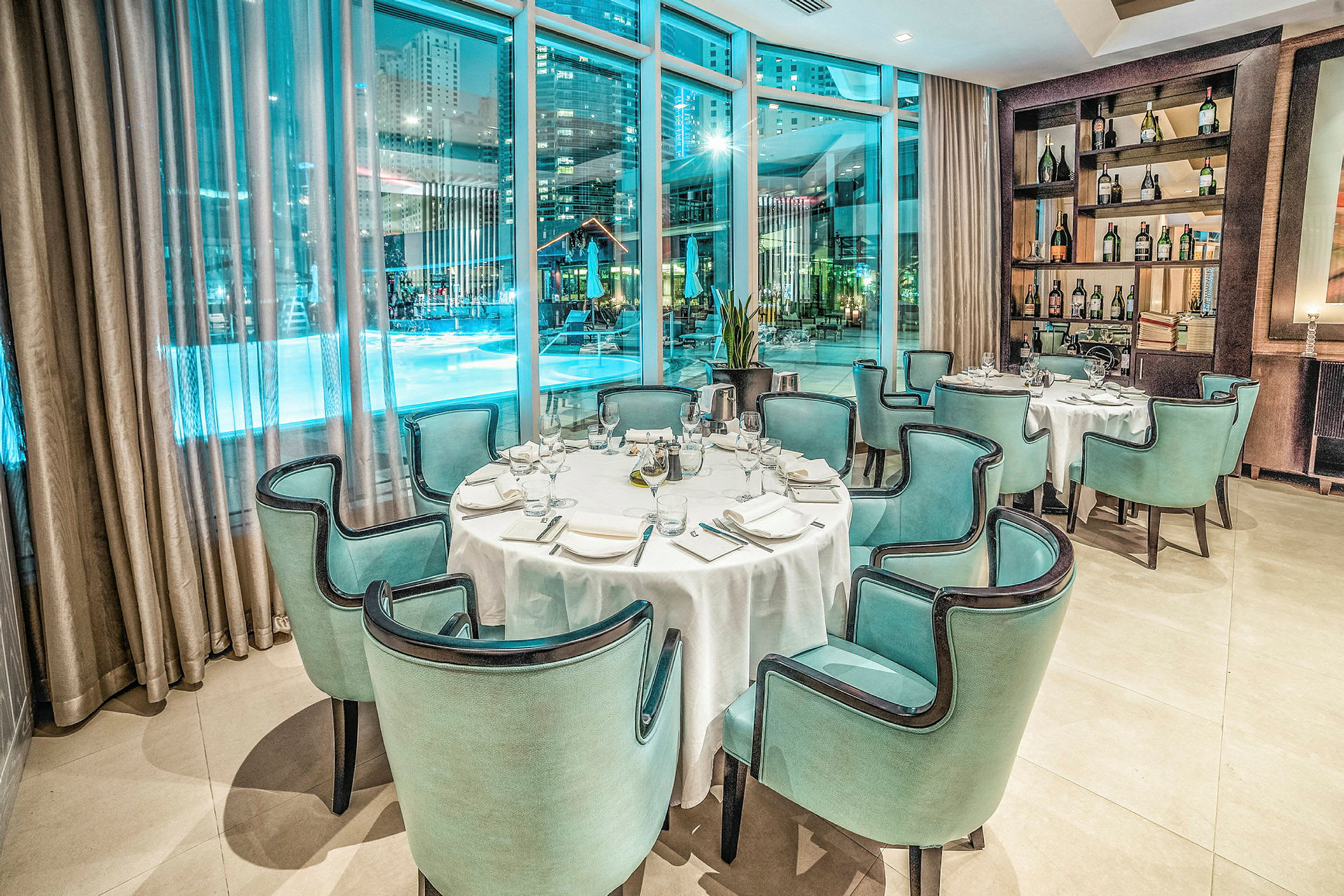 En elegant restaurant med opdækkede borde og grønblå stole, placeret ved store vinduer der giver udsigt over et oplyst poolområde om natten. (AI-genereret tekst)