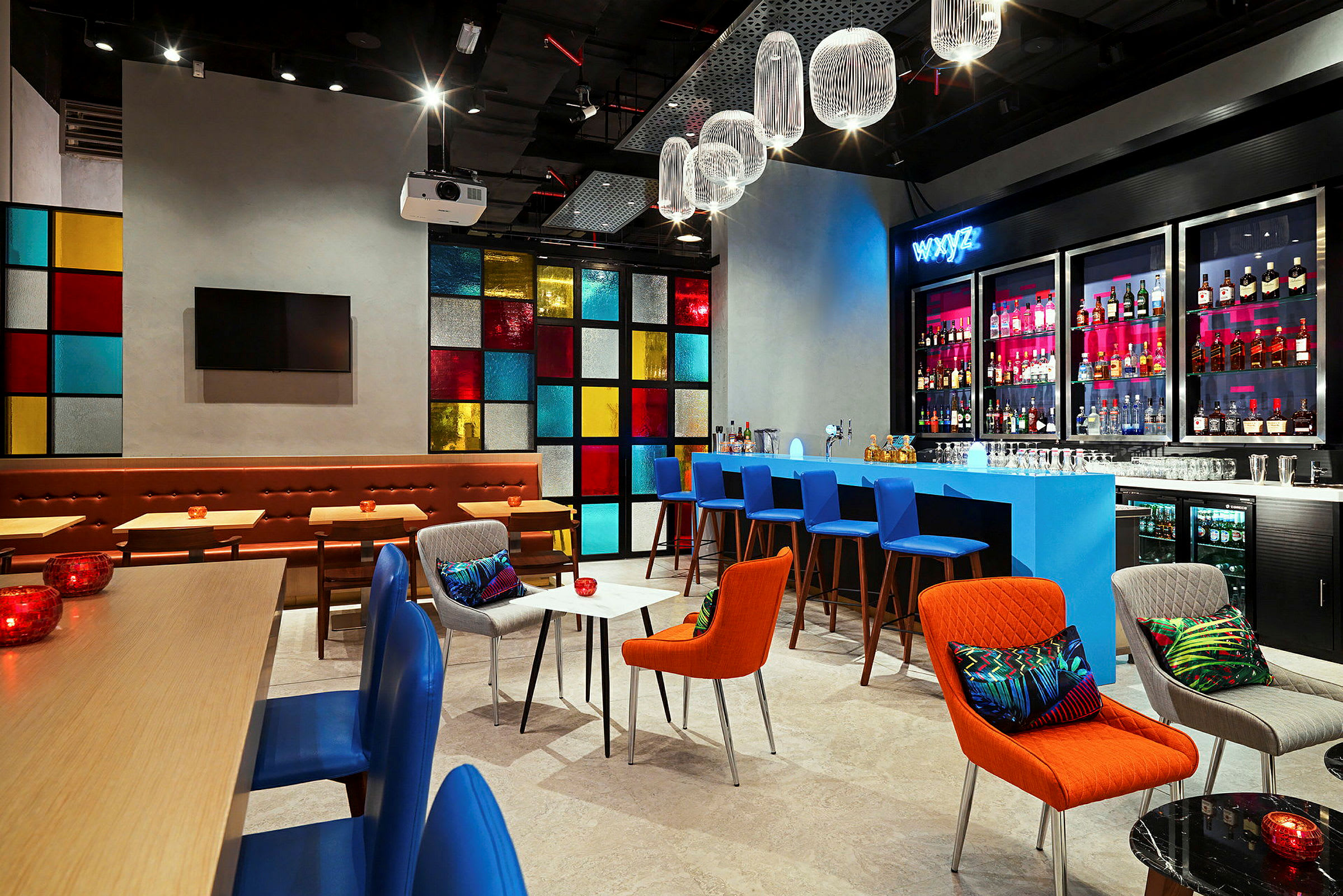 En moderne indrettet bar eller lounge med farverige møbler, en blå bardisk og hylder med flasker i baggrunden. Et vinduespanel med forskellige farvede ruder giver en livlig baggrund. Belysningen er varm, og designet er elegant og stilrent. (AI-genereret tekst)