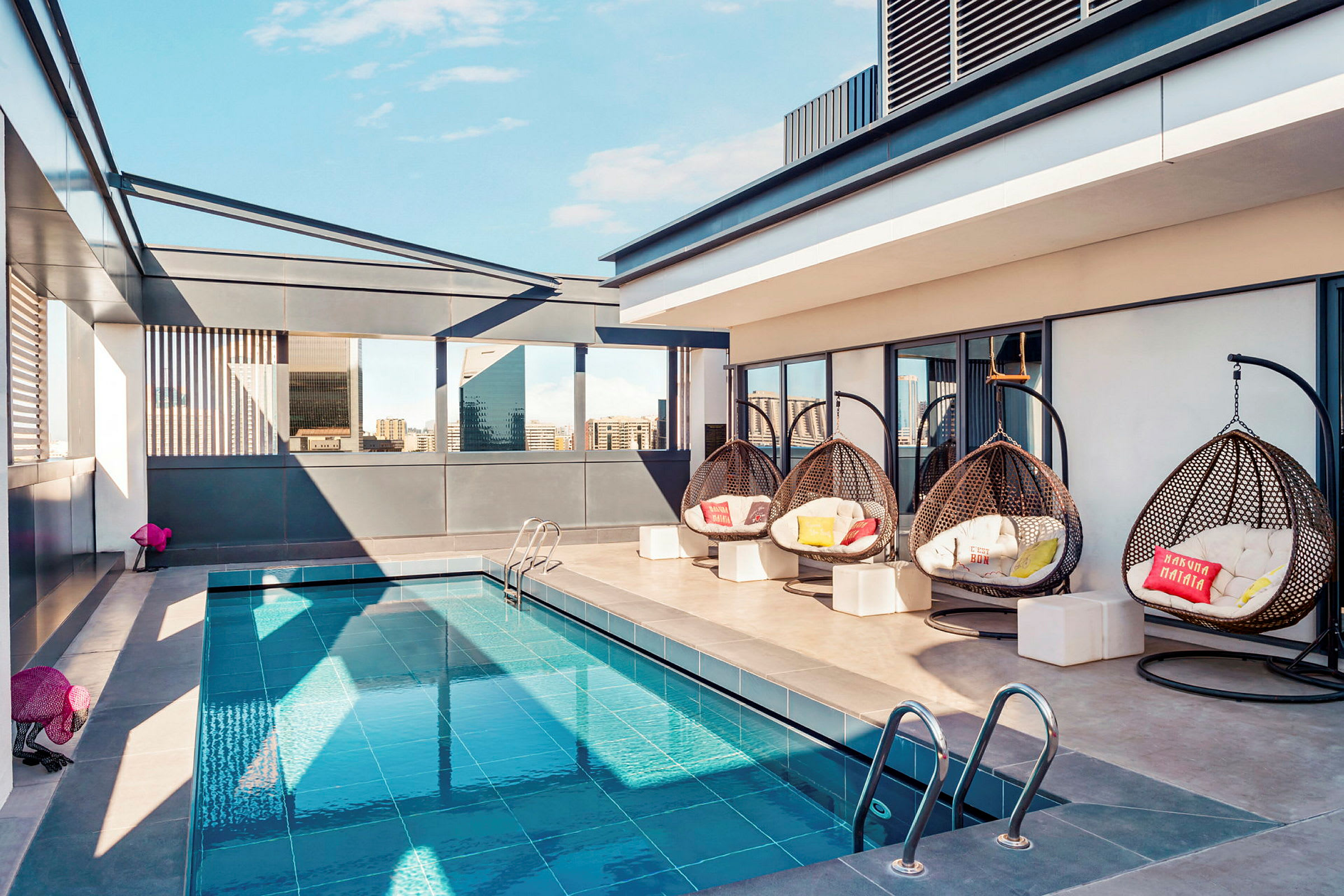 En tagterrasse med en lille pool, hængende flettede stole og moderne omgivelser. Bygninger ses i baggrunden. (AI-genereret tekst)