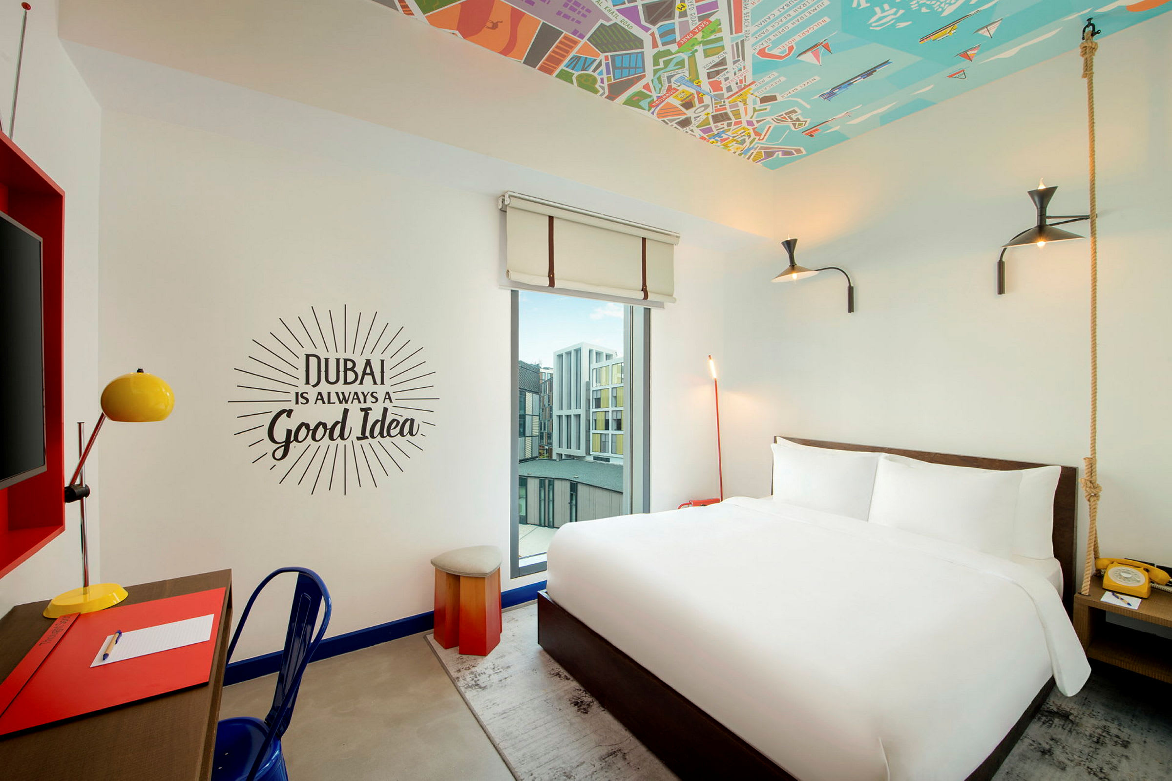 Et hotelværelse med en hvid dobbeltseng, skrivebord med lampe og en vægtekst, der siger 'Dubai is always a good idea'. (AI-genereret tekst)
