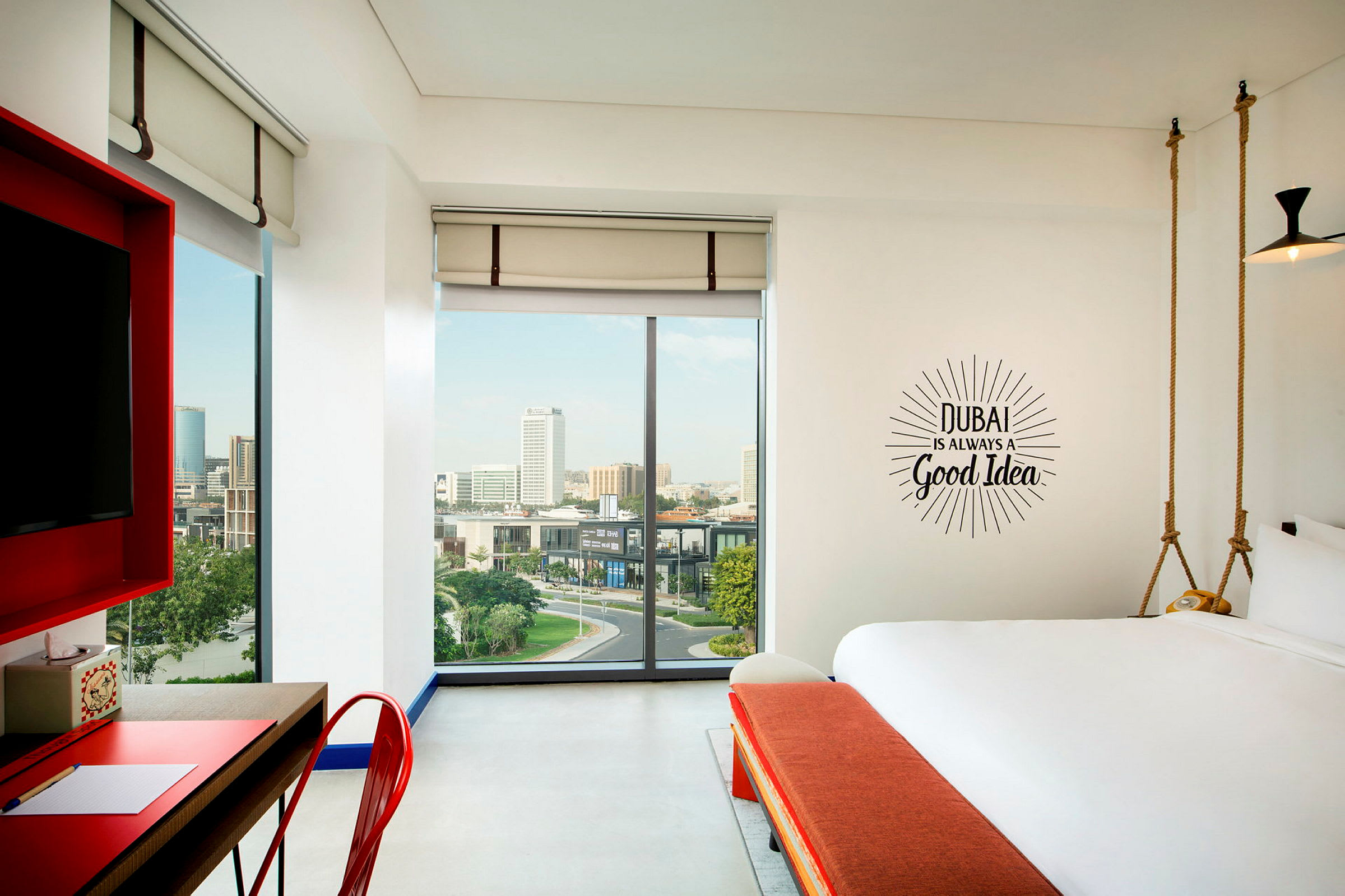 Et moderne hotelværelse med store vinduer og udsigt over en by. På væggen står teksten: "Dubai is always a good idea". (AI-genereret tekst)
