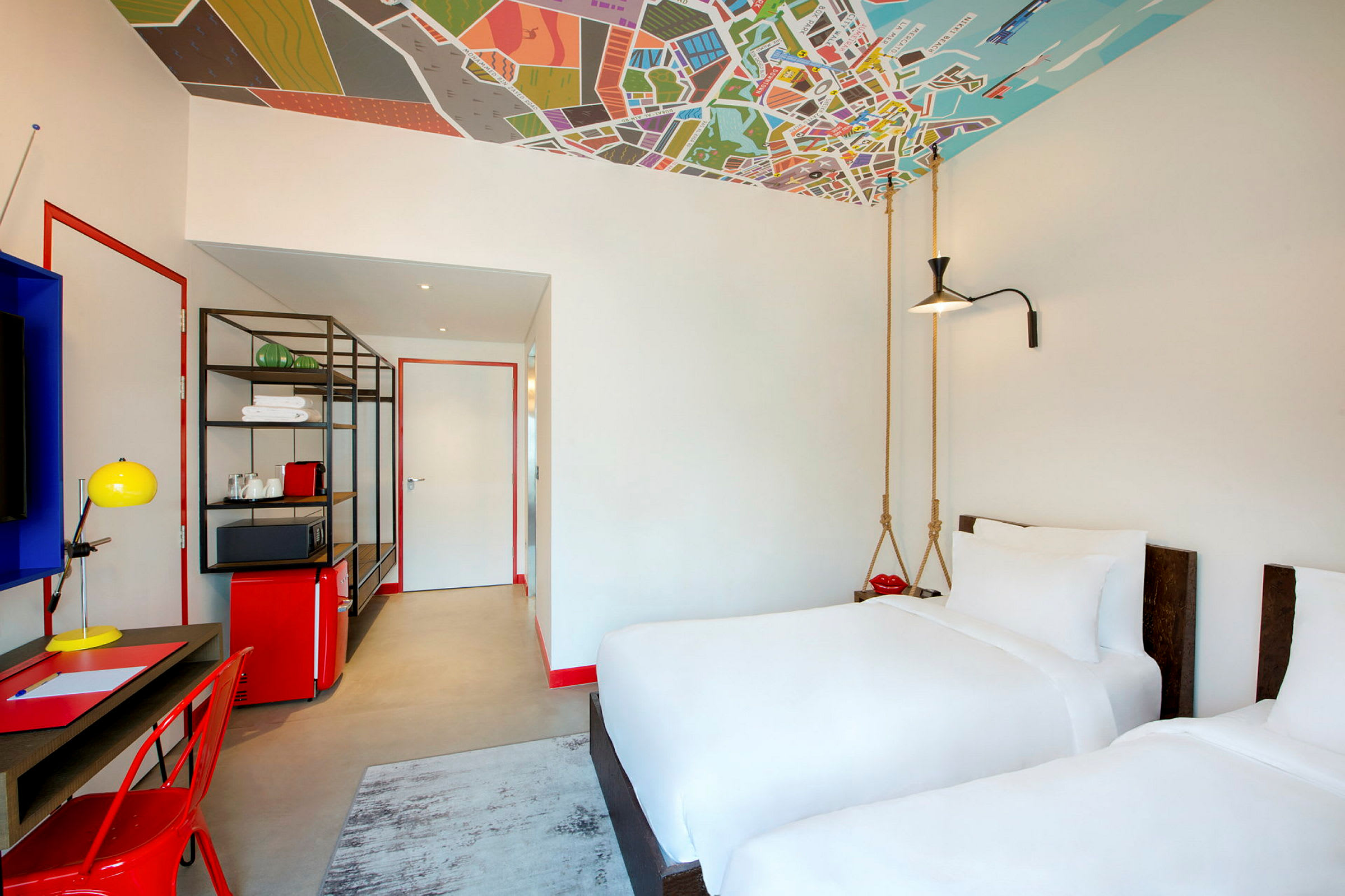 Et moderne hotelværelse med to hvide enkeltsenge, et skrivebord, en rød stol og farverige indretningsdetaljer. Loftet er dekoreret med et kortmønster. (AI-genereret tekst)