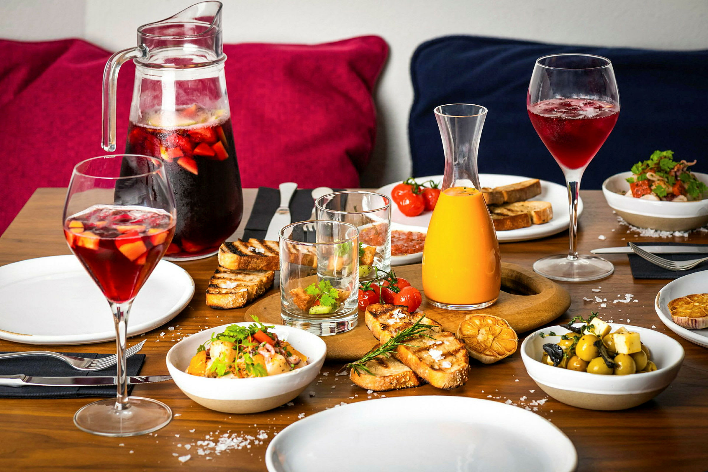 Et dækket bord med forskellige retter og drikkevarer, herunder brød, salat, oliven og kander med juice og røde drikke. (AI-genereret tekst)