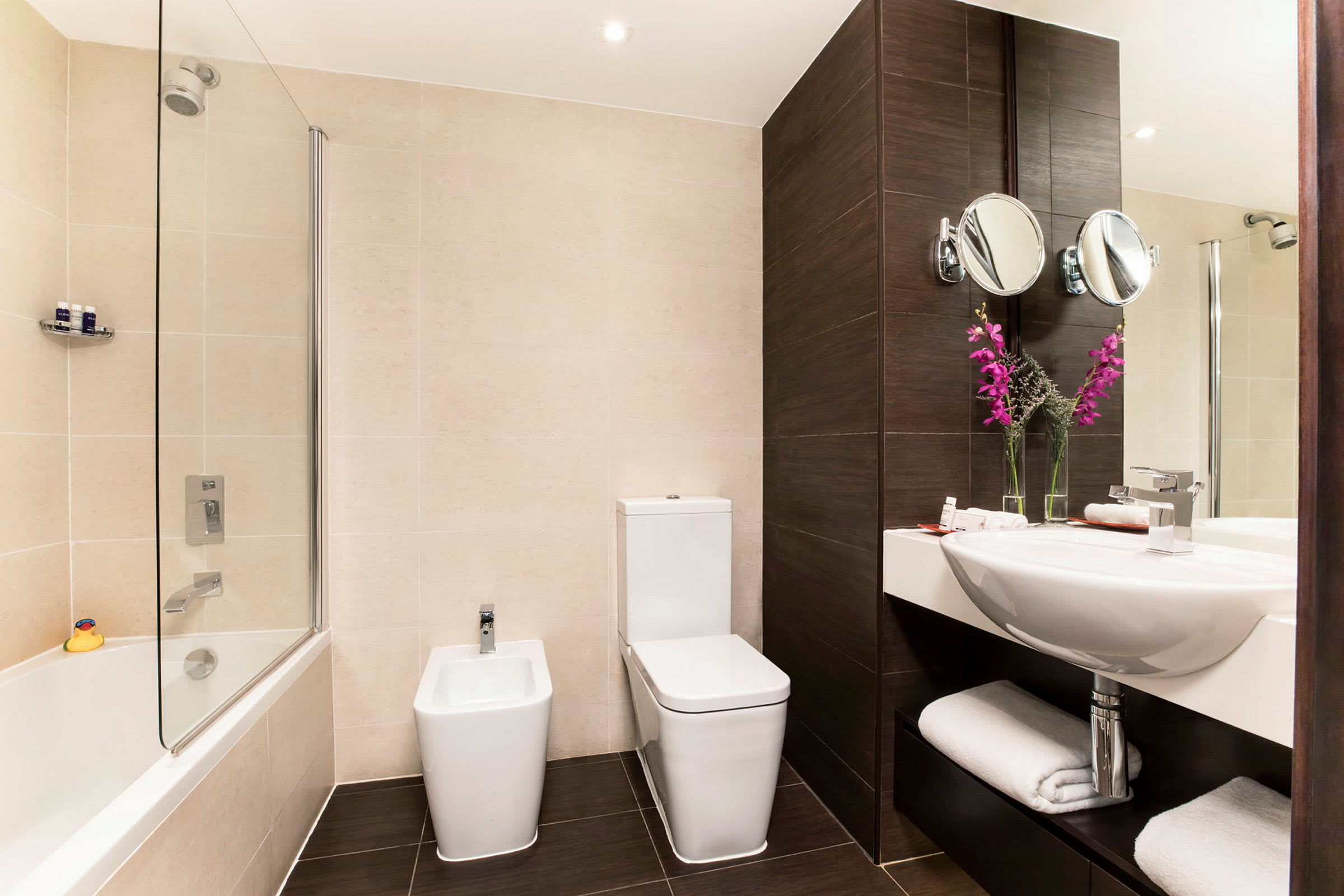 Et moderne badeværelse med et badekar, et toilet, et bidet og en håndvask med spejl. Dekorationen omfatter lyserøde blomster i glasvaser. (AI-genereret tekst)