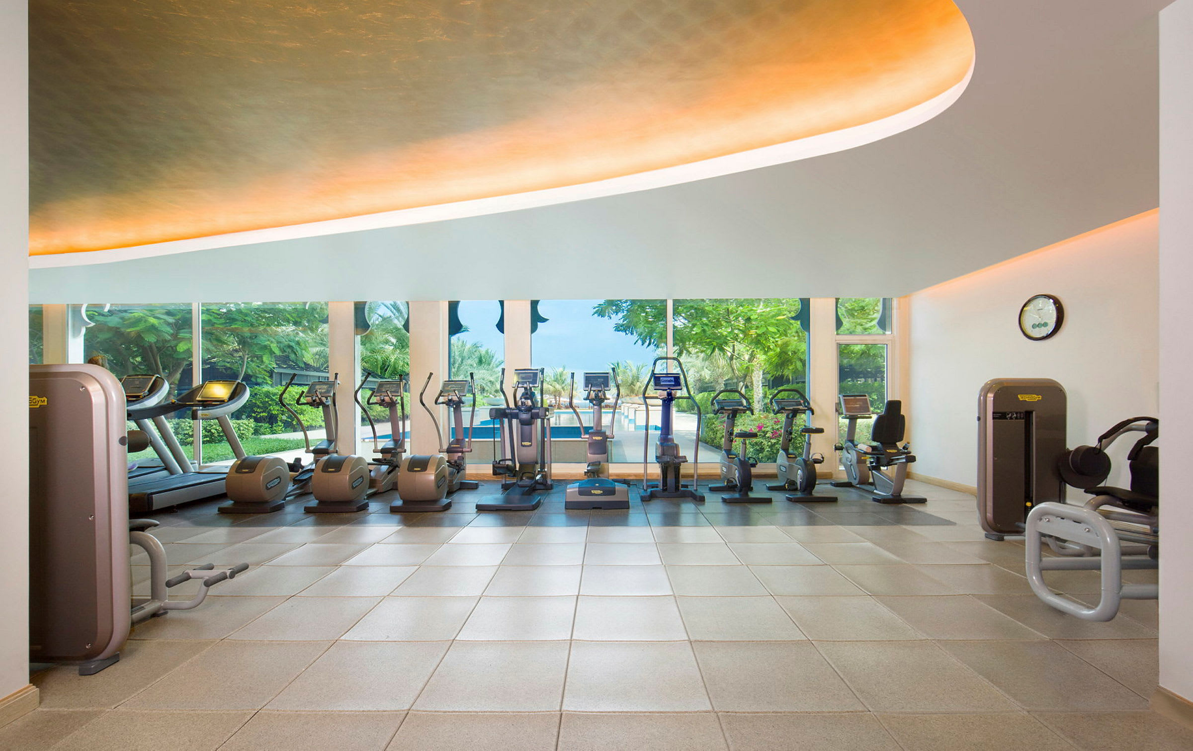 Et fitnesscenter med træningsmaskiner og vinduer med udsigt til have og poolområde. (AI-genereret tekst)