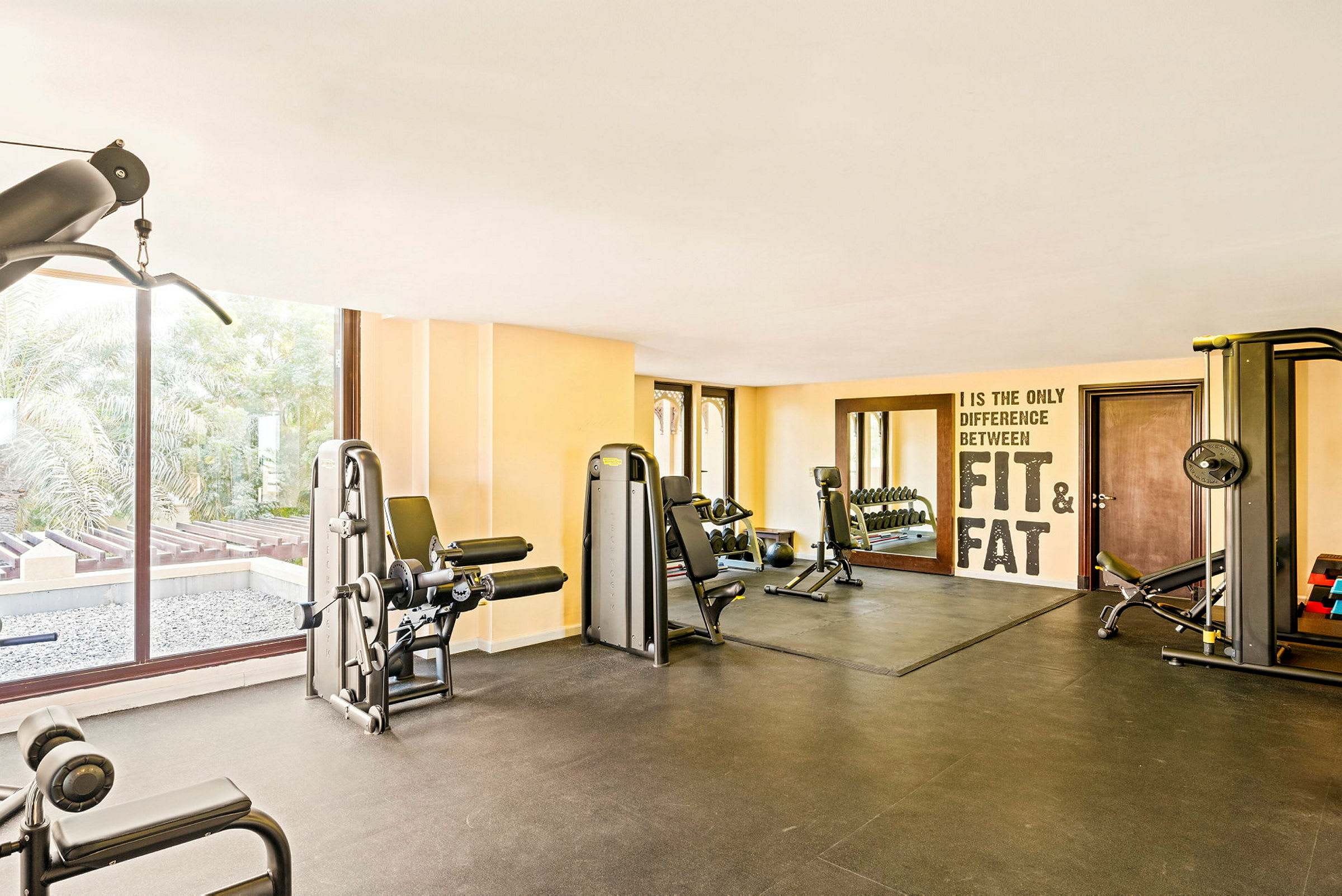 Et motionsrum med fitnessmaskiner, en spejlvæg og vinduer med udsigt til grønne omgivelser. (AI-genereret tekst)