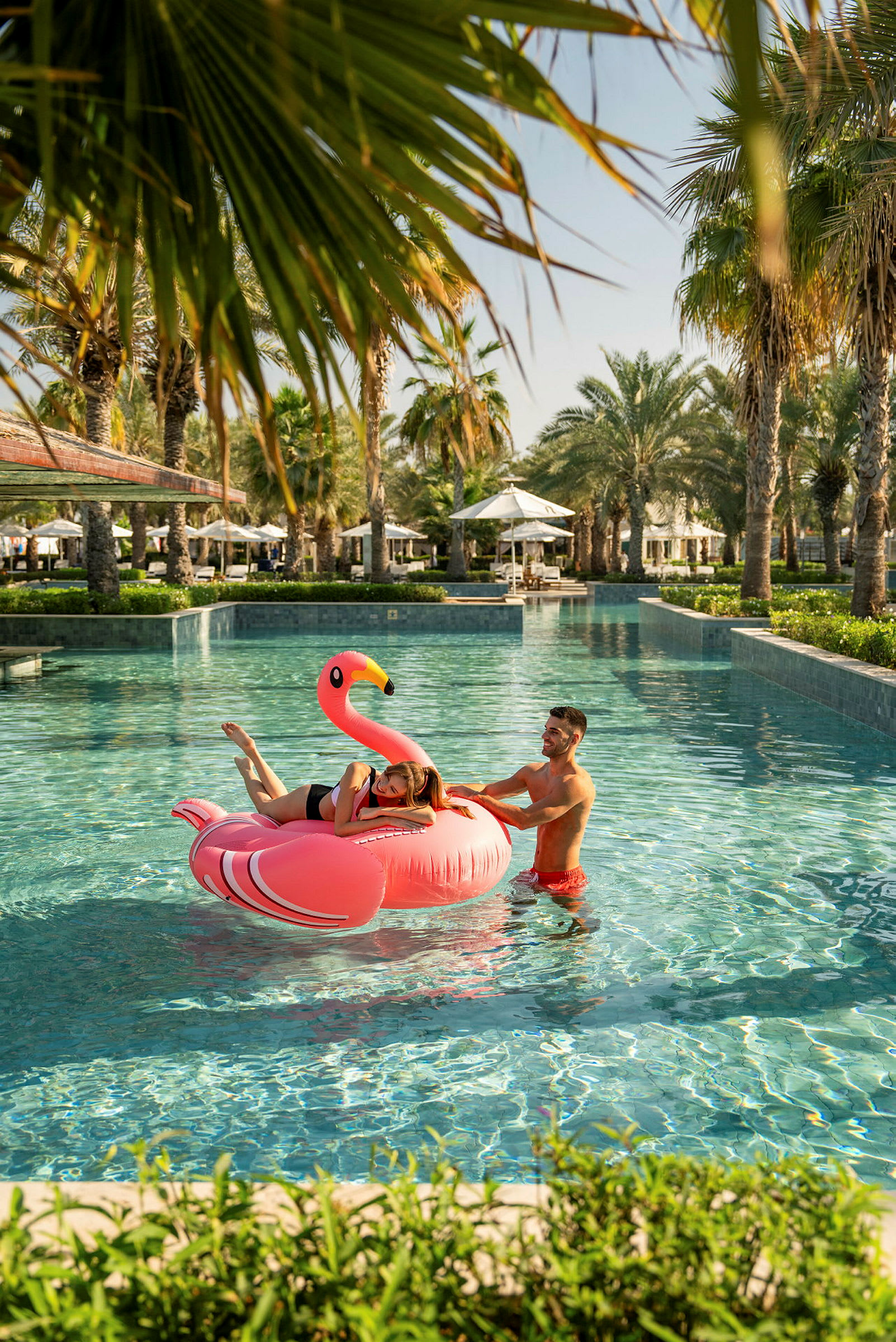 To personer ved en swimmingpool med en oppustelig flamingo. Palme- og liggestole ses i baggrunden. (AI-genereret tekst)