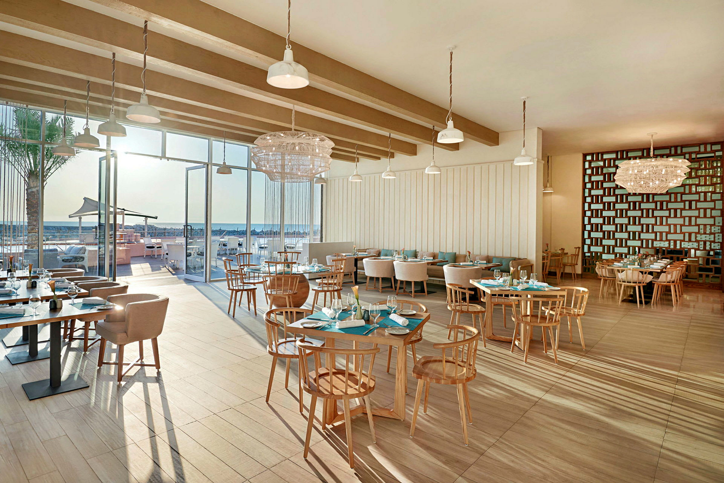 En moderne restaurant indendørs med store glasvinduer ud mod en udendørs servering og stranden. (AI-genereret tekst)