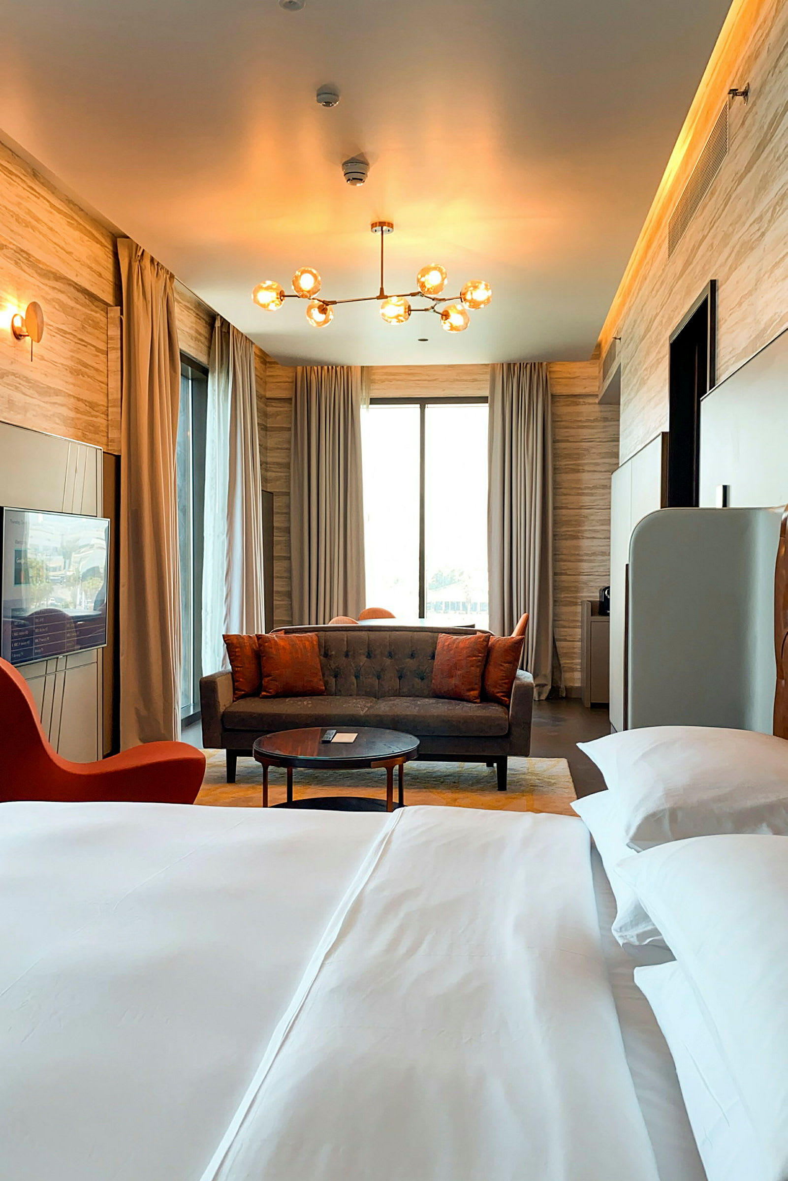 Et elegant møbleret hotelværelse med en stor seng i forgrunden og en sofagruppe foran et stort vindue med gardiner. En moderne lampe hænger i loftet. (AI-genereret tekst)