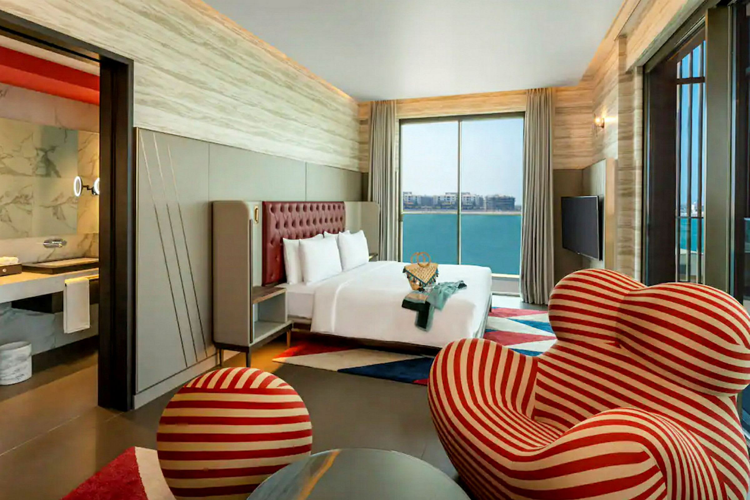 Et moderne hotelværelse med en stor seng placeret foran et stort vindue med udsigt over havet. Værelset har også en unik stribet rød og hvid lænestol med en matchende fodskammel. (AI-genereret tekst)