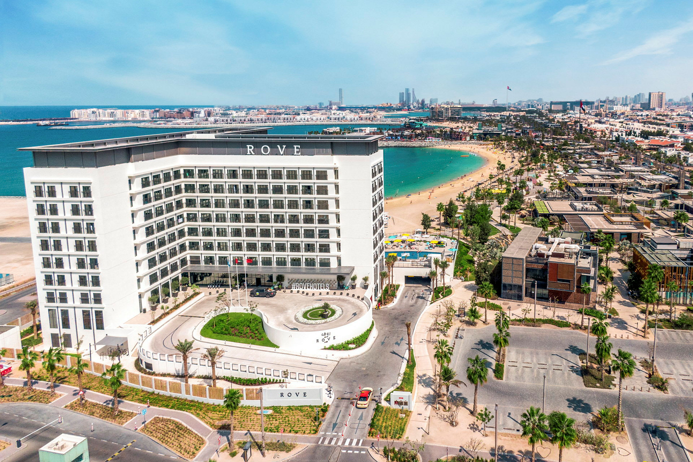 Et hotel med en hvid facade ved en strand med blåt vand. (AI-genereret tekst)