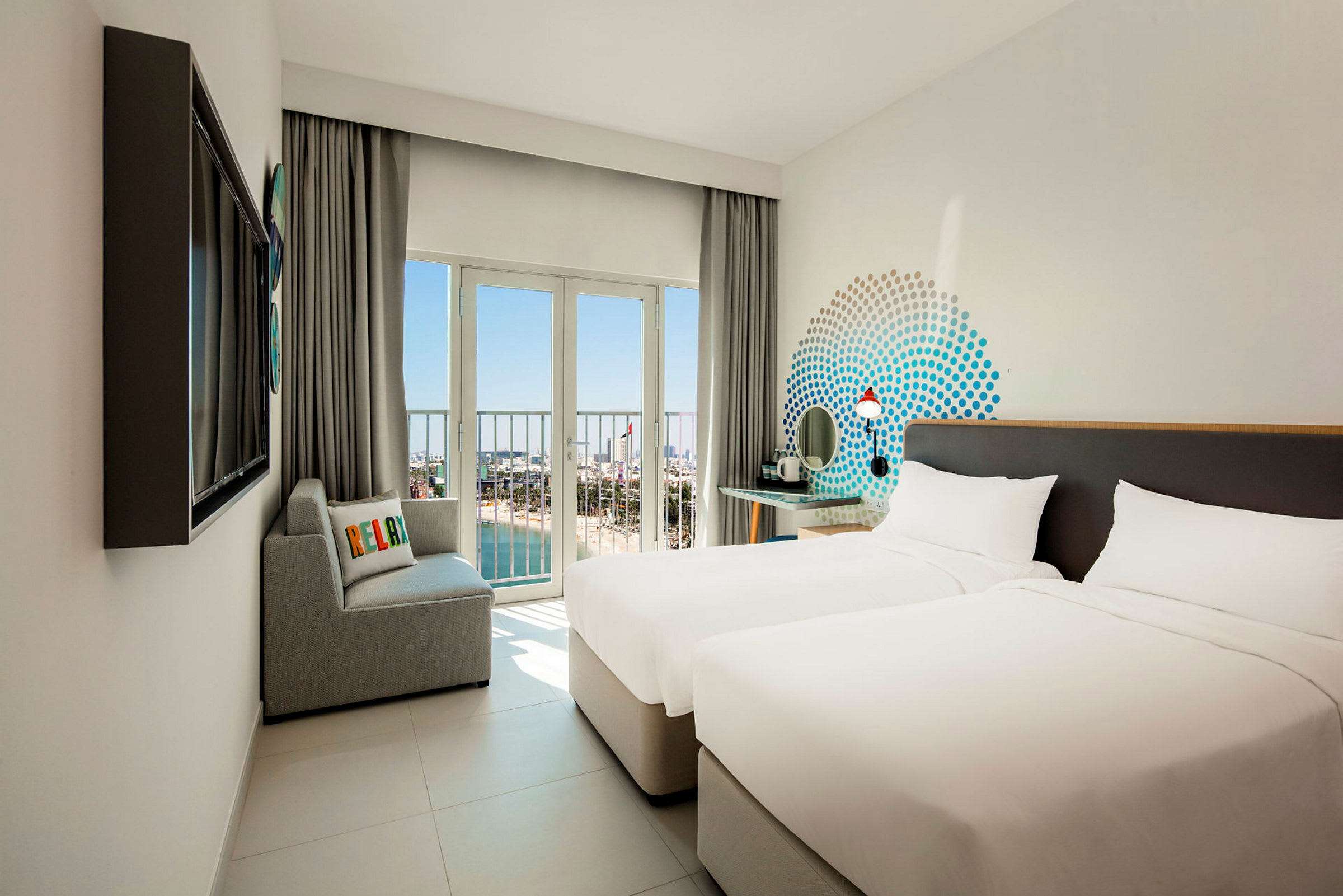 Hotelværelse med to enkeltsenge, en sofa og udsigt gennem store vinduer mod stranden og havet. (AI-genereret tekst)