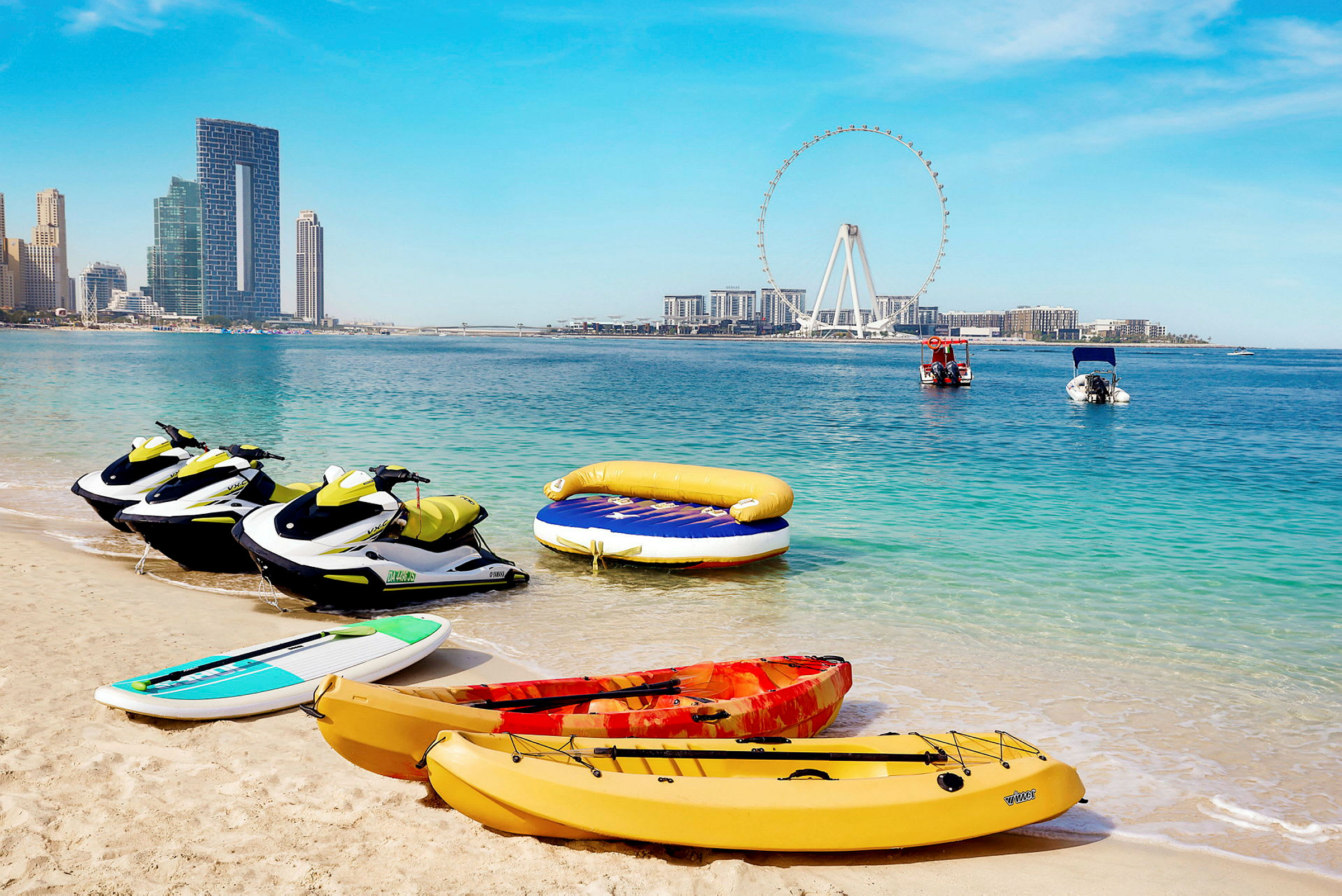 Jetski, kajakker og et paddleboard på stranden med havet og byens skyline i baggrunden. (AI-genereret tekst)