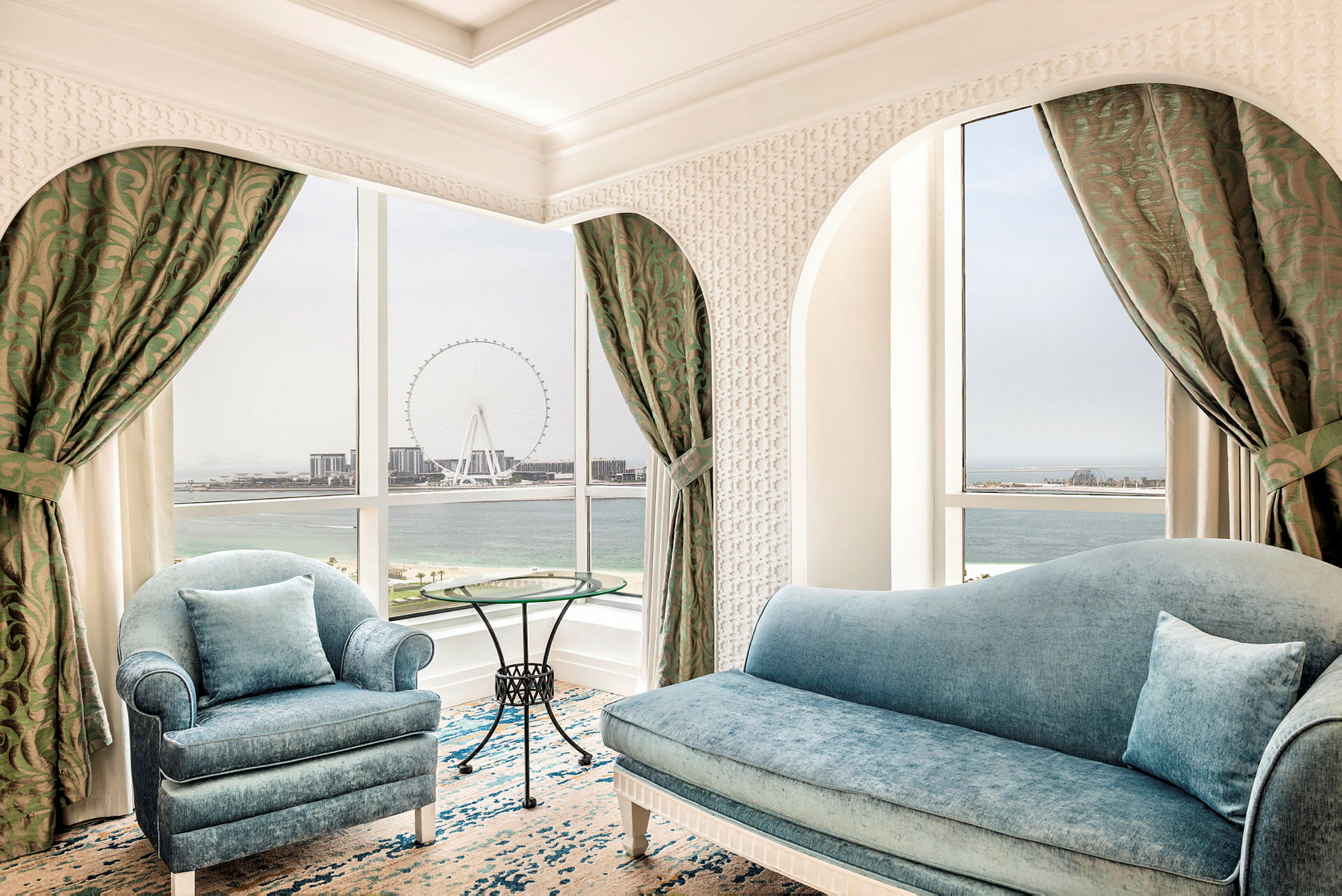 Et elegant værelse med blå sofaer og et rundt glasbord. Vinduerne giver udsigt over havet og en stor Pariserhjul. (AI-genereret tekst)