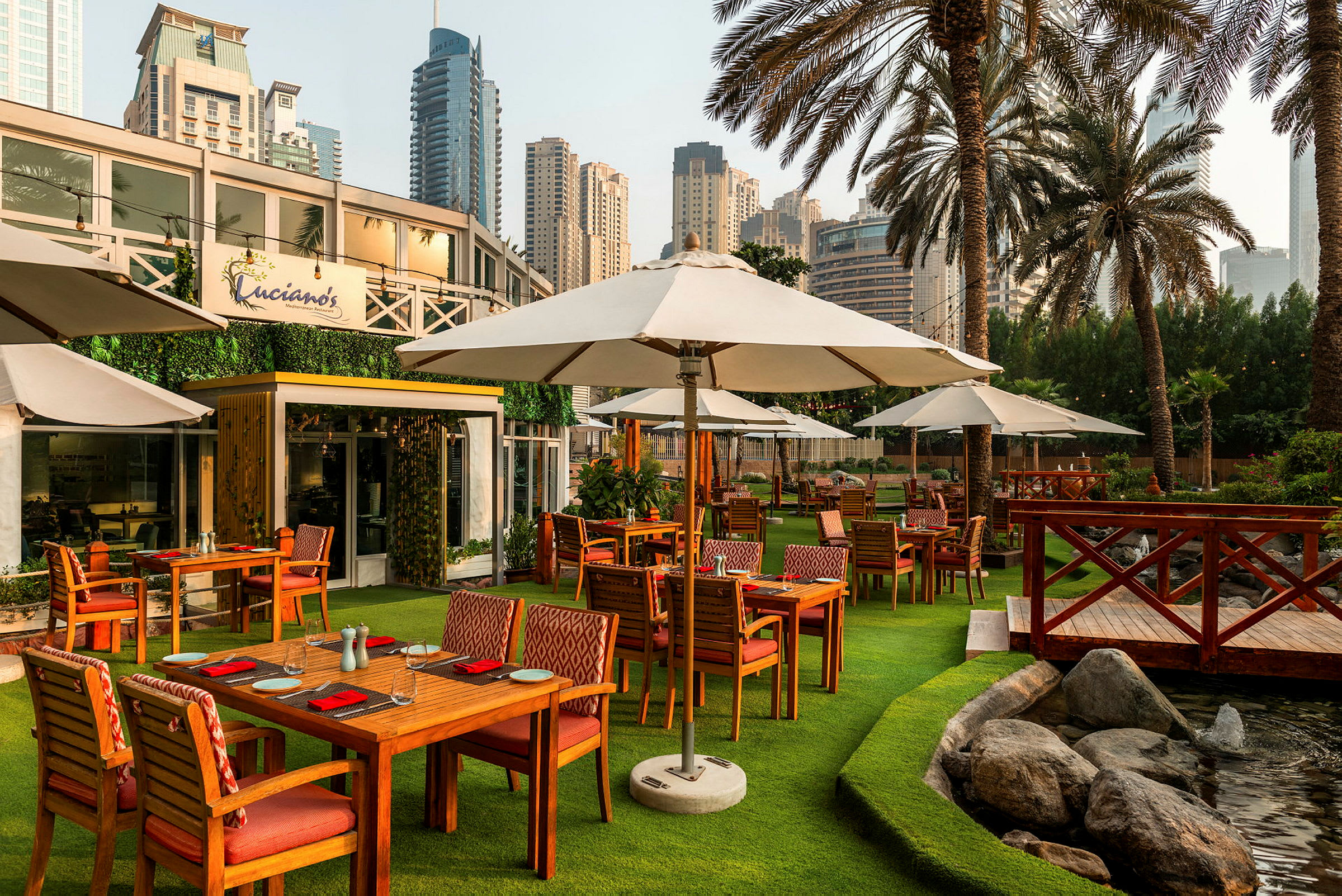 Udendørs miljø med restaurantborde, parasoller og grønt græs, omgivet af palmer og byens skyline. (AI-genereret tekst)