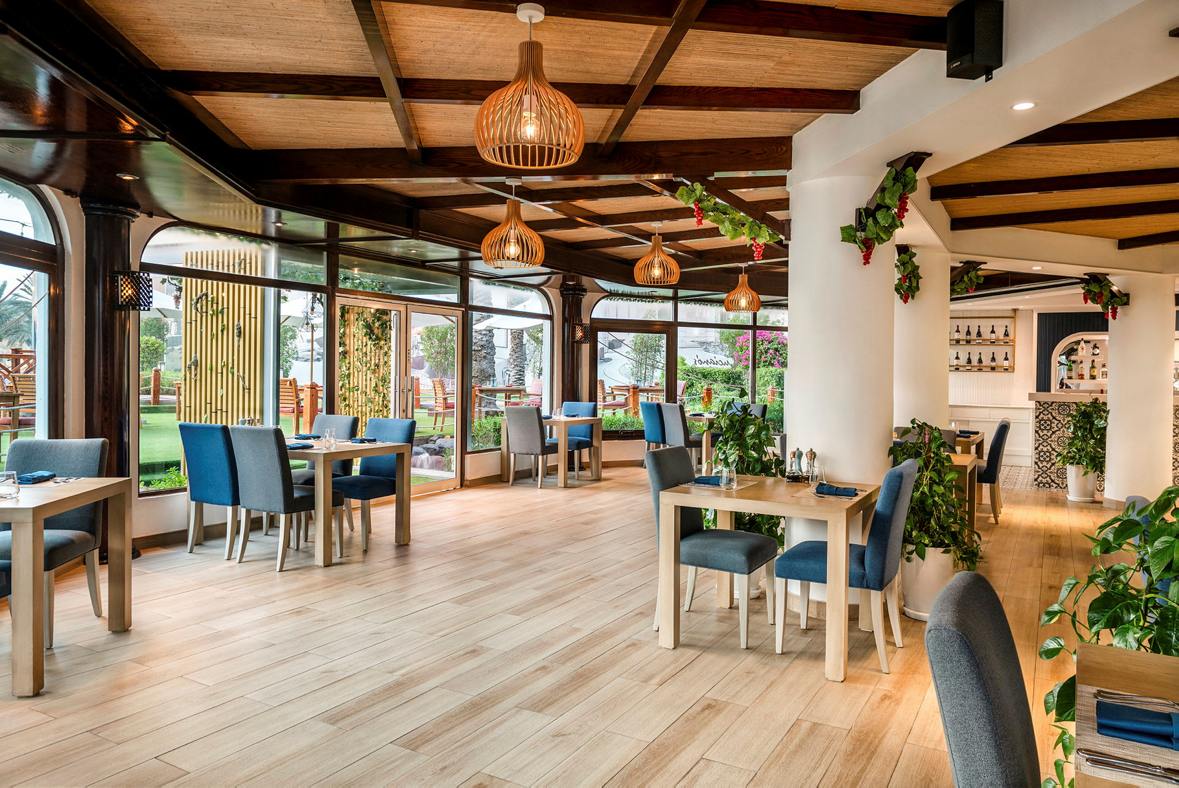 En lys restaurant med borde og stole i moderne trædesign, grønne planter og store vinduespartier med udsigt til en have. (AI-genereret tekst)