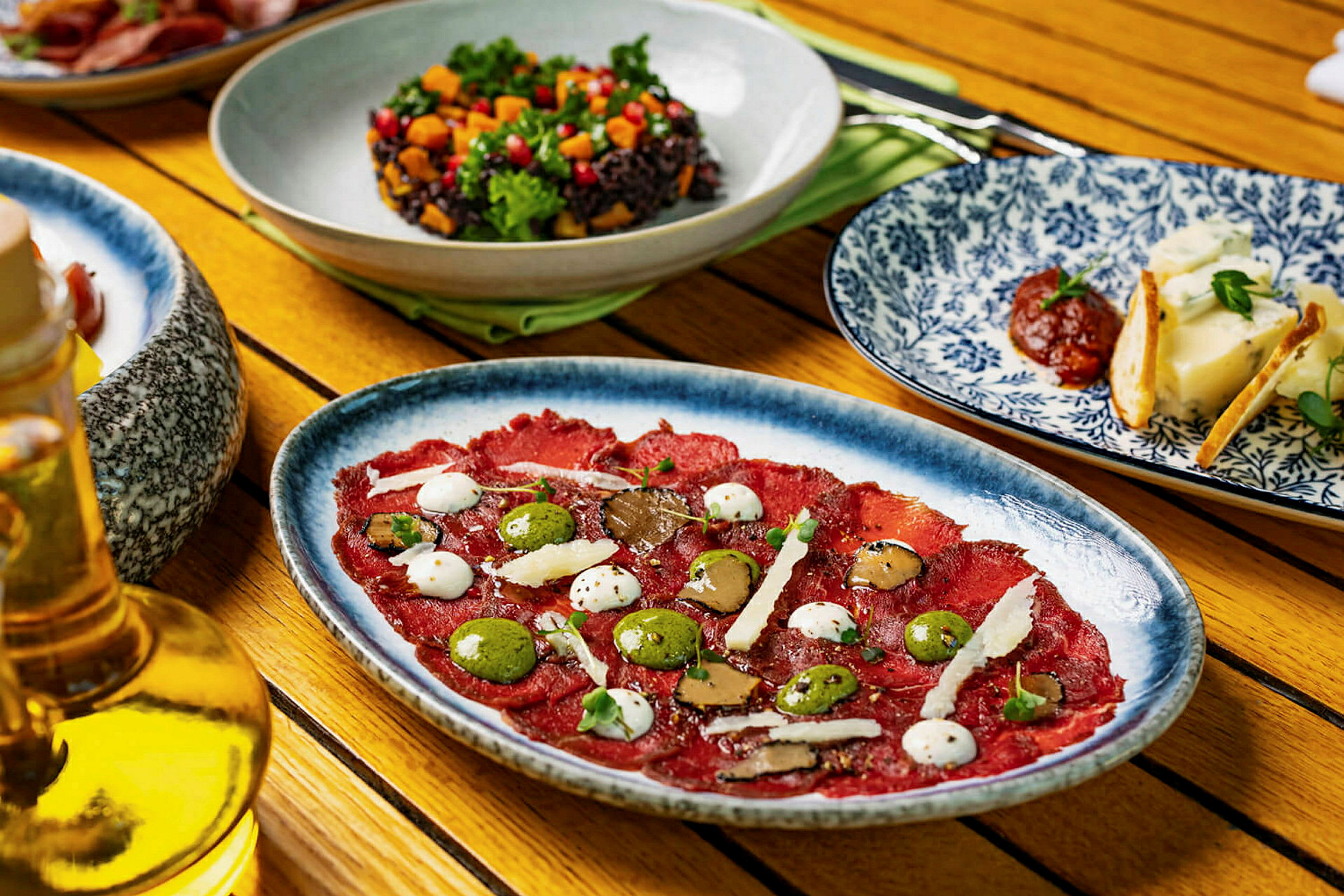 Et bord med forskellige retter, herunder en tallerken med carpaccio, en skål med salat og tallerkener med ost og tilbehør. (AI-genereret tekst)
