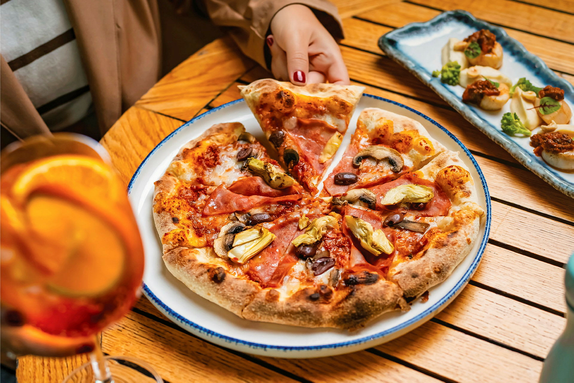 En tallerken med pizza toppet med skinke, oliven, svampe og artiskokker på et bord. En person tager et pizzastykke. Bag pizzaen ses et fad med små forretter og ved siden af et glas med orange drik. (AI-genereret tekst)