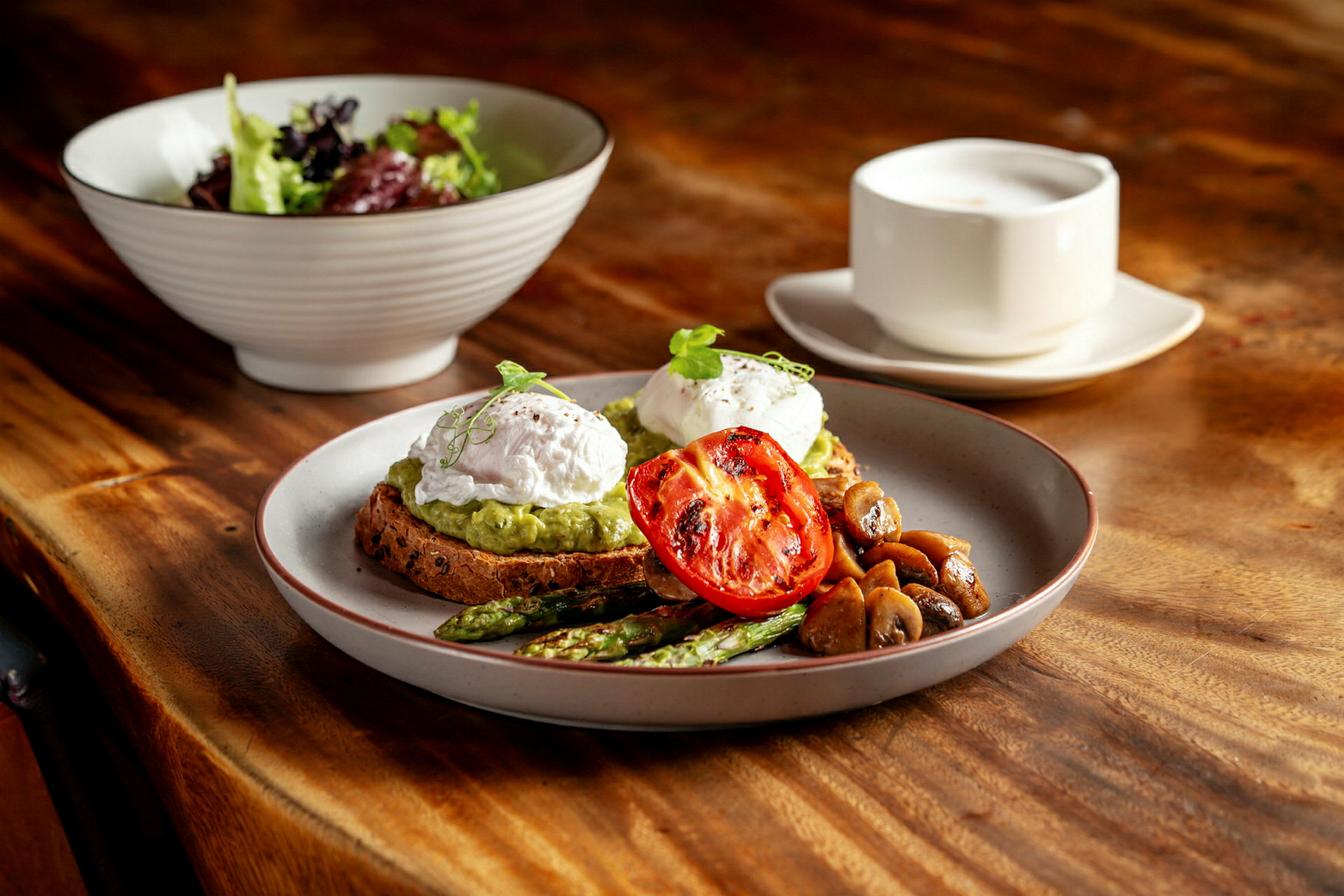En tallerken med toast, moset avocado, pocherede æg, grillet tomat, asparges og svampe. Bagved ses en salatskål og en kop på et træbord. (AI-genereret tekst)