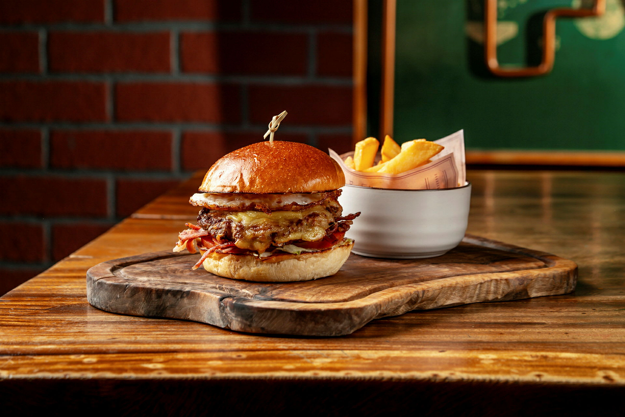 En burger med brød, kød og ost serveret på en træplade. Ved siden af står en hvid skål med pommes frites. (AI-genereret tekst)