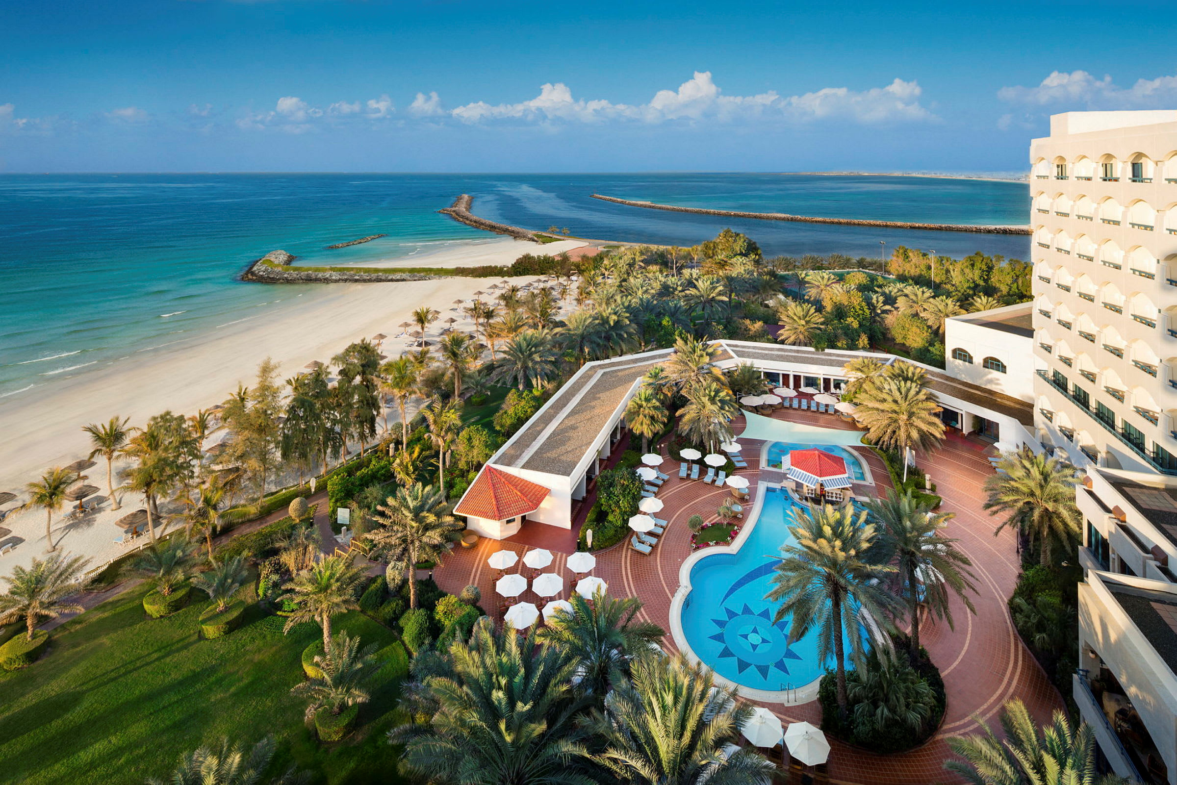 Et billede af et hotel ved stranden med en udendørs pool, liggestole og omkringliggende beplantning. (AI-genereret tekst)