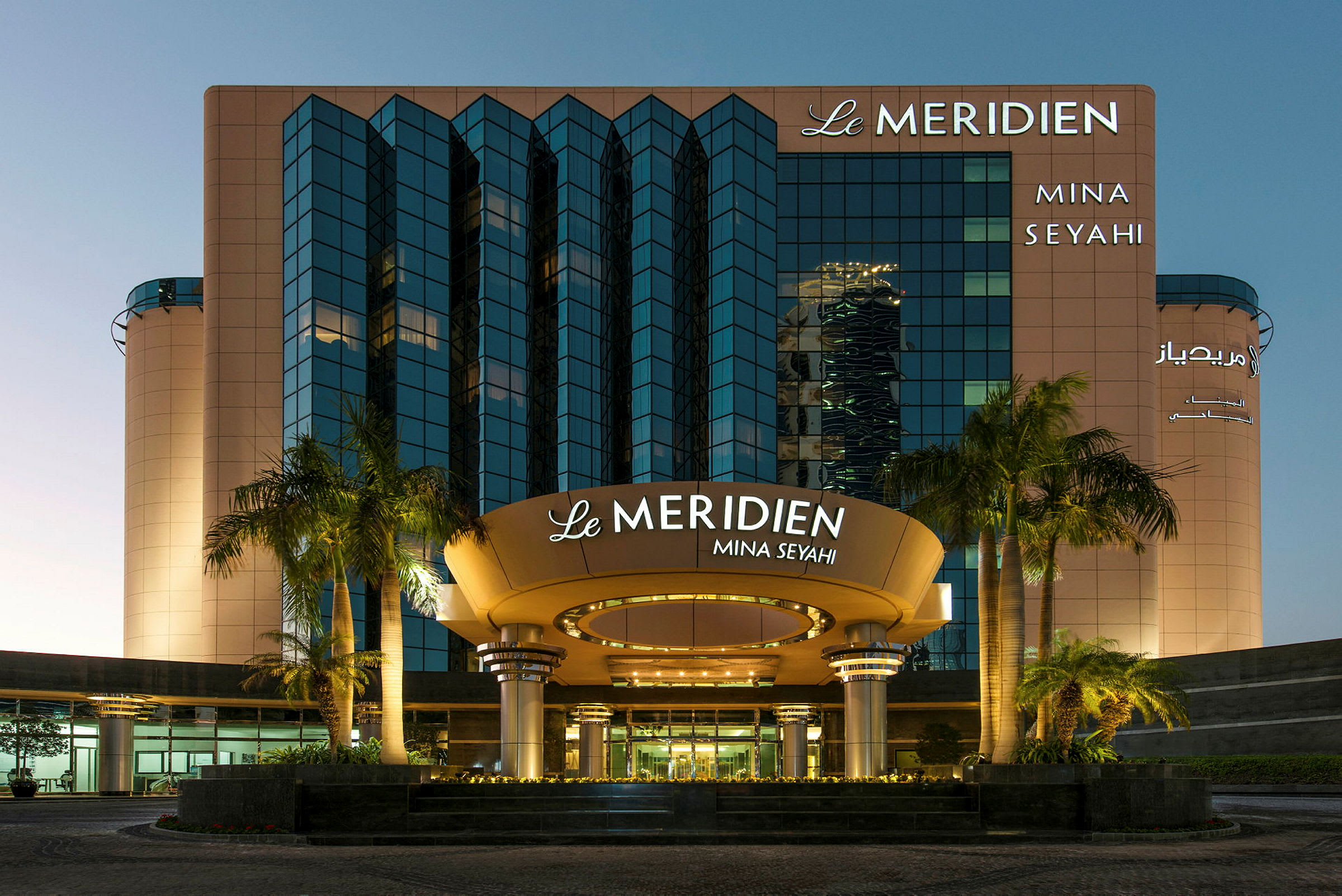 Facaden af Le Meridien Mina Seyahi hotel med palmer i forgrunden. (AI-genereret tekst)
