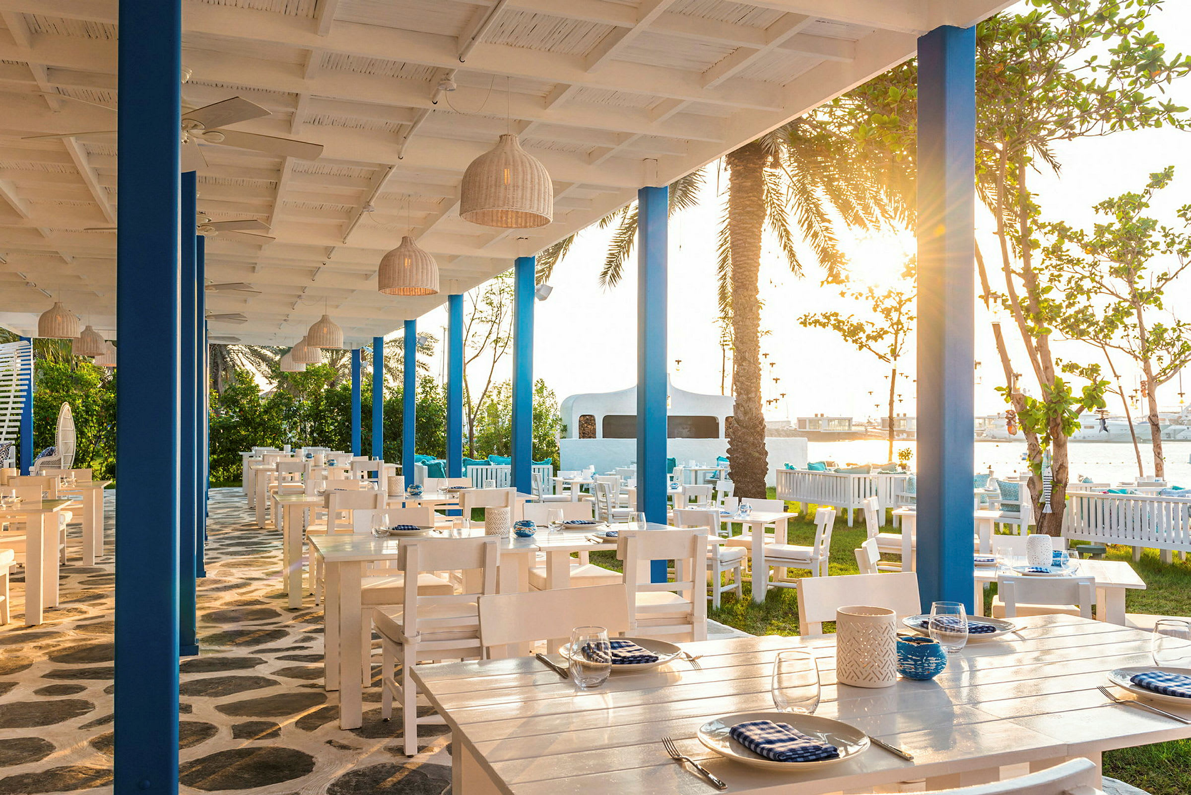 Restaurant med hvide møbler og blå detaljer, udendørs med solnedgang i baggrunden. (AI-genereret tekst)