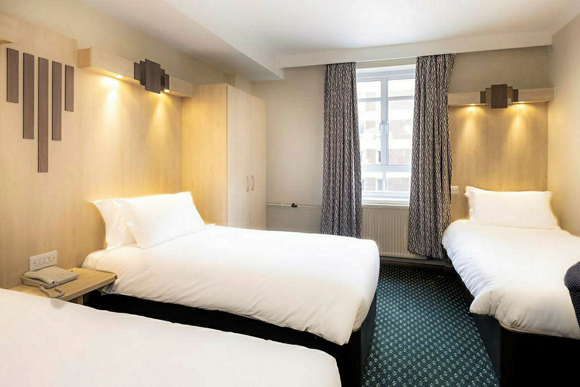 Et hotelværelse med tre senge, gardiner og et vindue. (AI-genereret tekst)