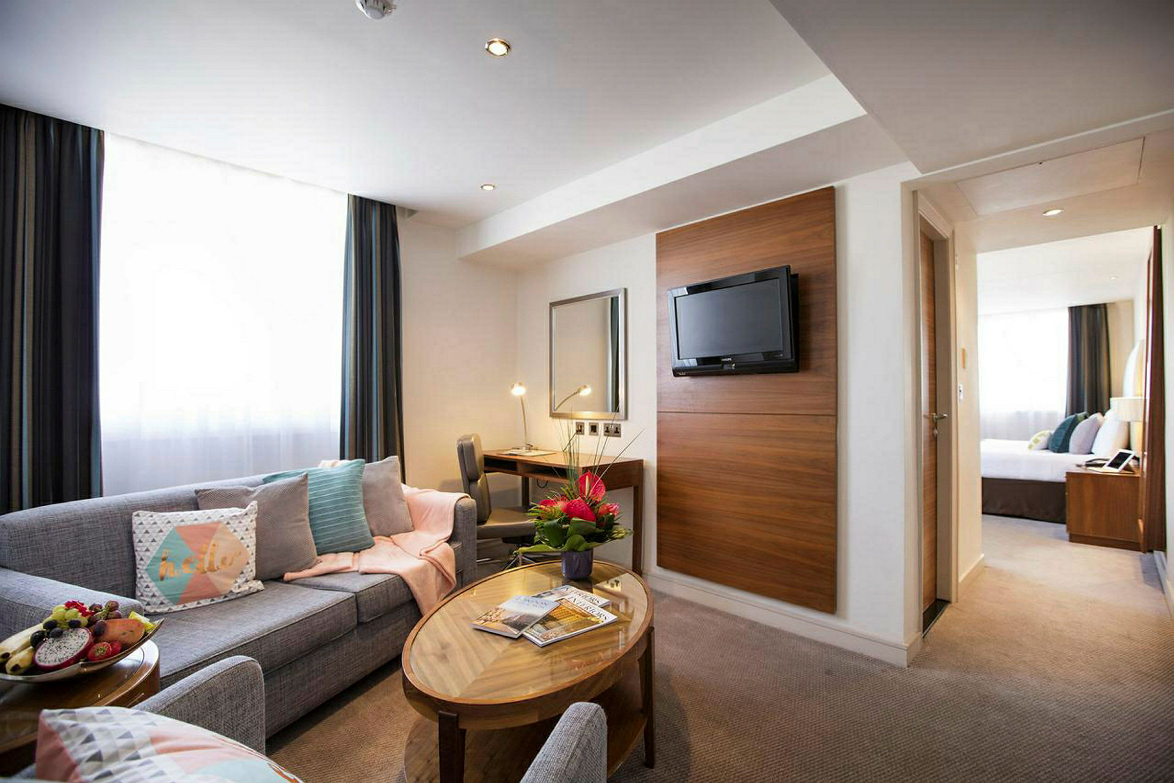 Et hotelværelse med sofagruppe, sofabord, skrivebord, vægmonteret TV og blomster. (AI-genereret tekst)