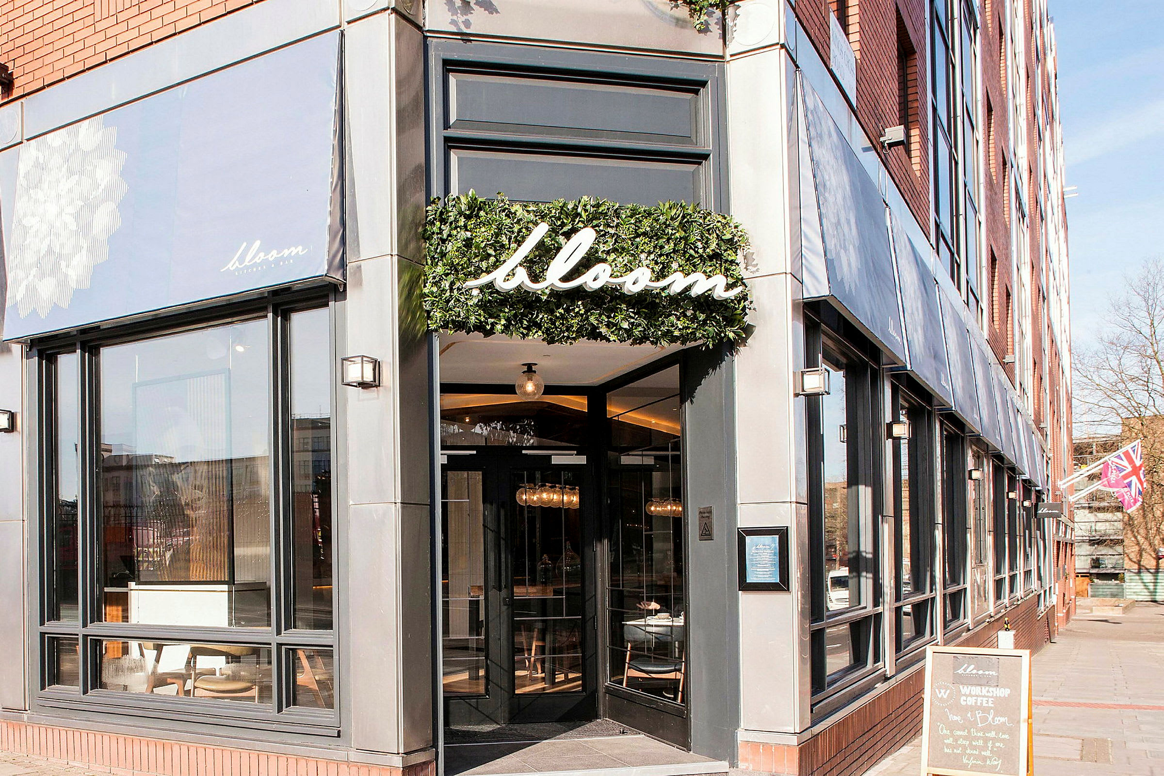 Indgangen til restauranten Bloom med store vinduer og et skilt med navnet omkranset af grønt. (AI-genereret tekst)