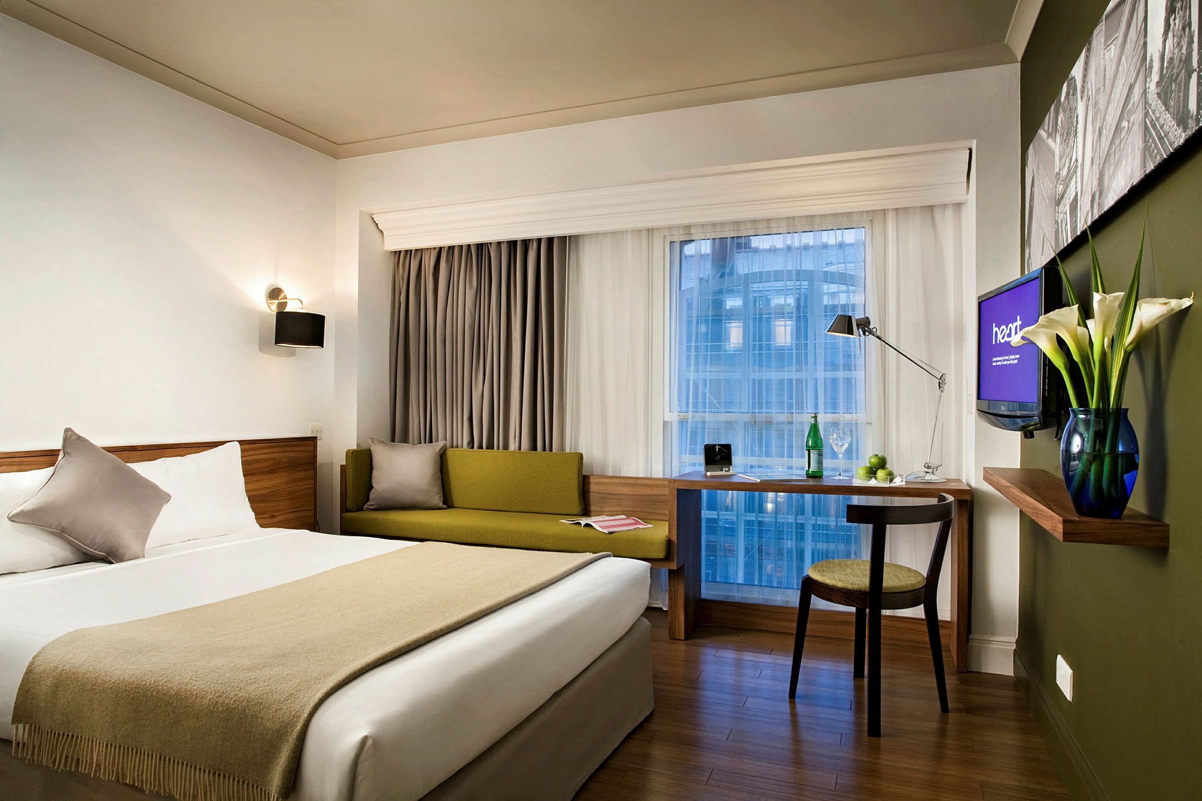 Et moderne hotelværelse med en dobbeltseng, grøn sofa, skrivebord og stort vinduesudsyn. (AI-genereret tekst)