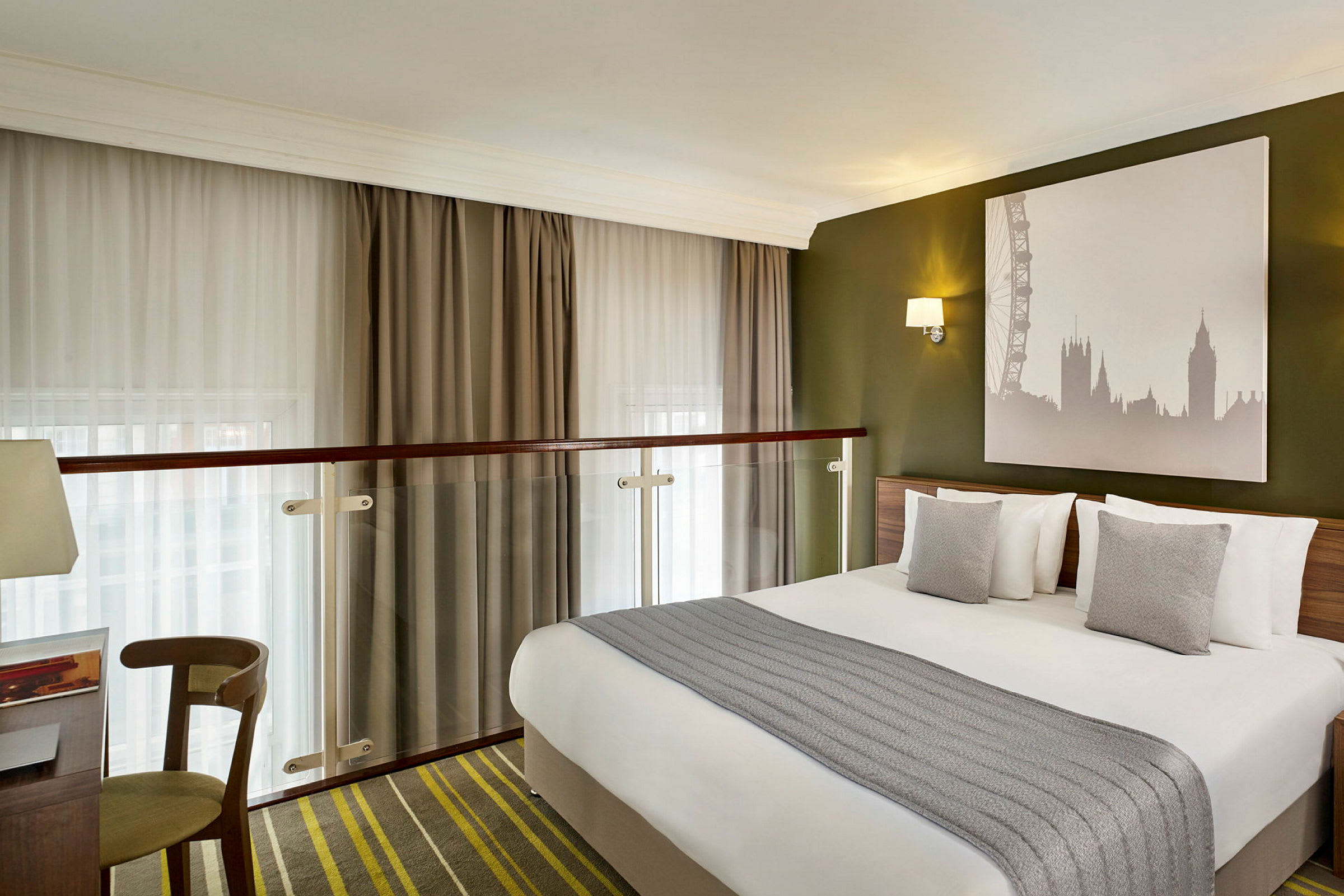 Et hotelværelse med en dobbeltseng, grå puder og et billede af Londons skyline på væggen. (AI-genereret tekst)