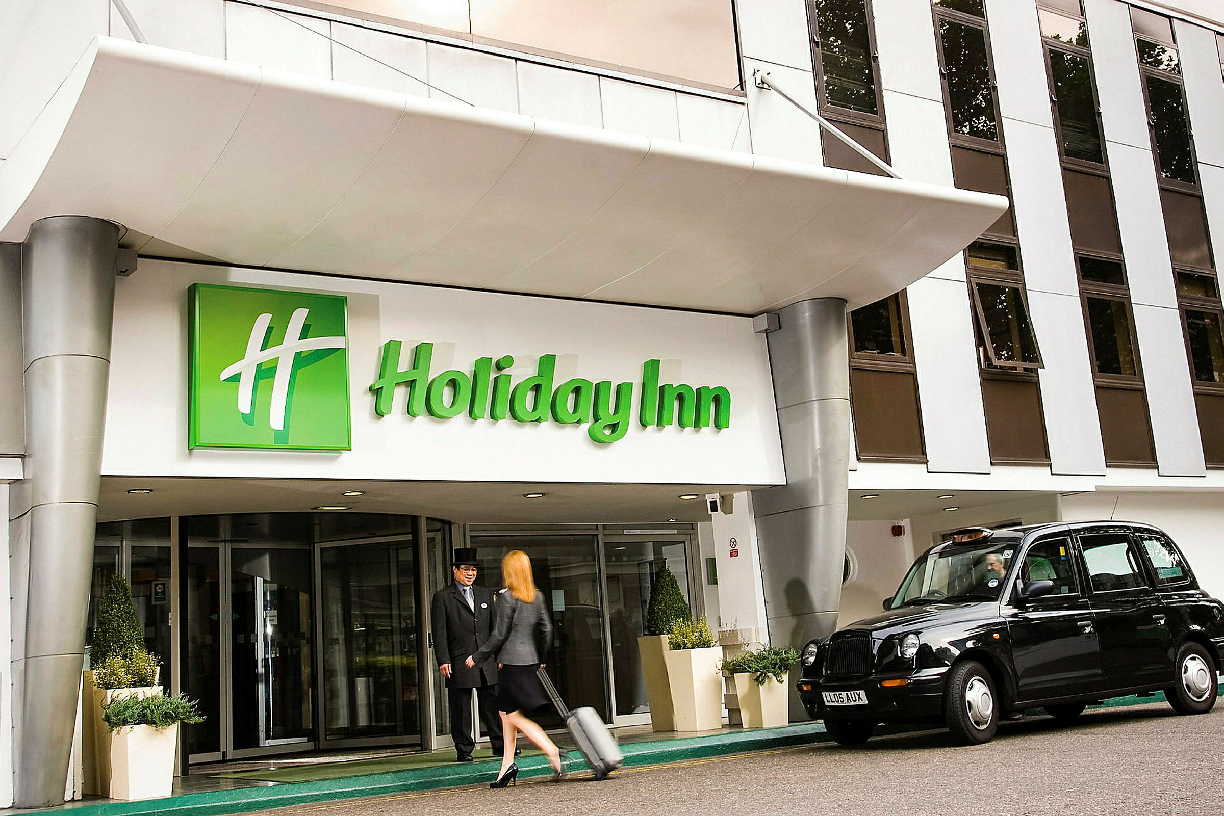 Indgangen til Holiday Inn med en taxa parkeret udenfor og en person, der trækker en kuffert. (AI-genereret tekst)