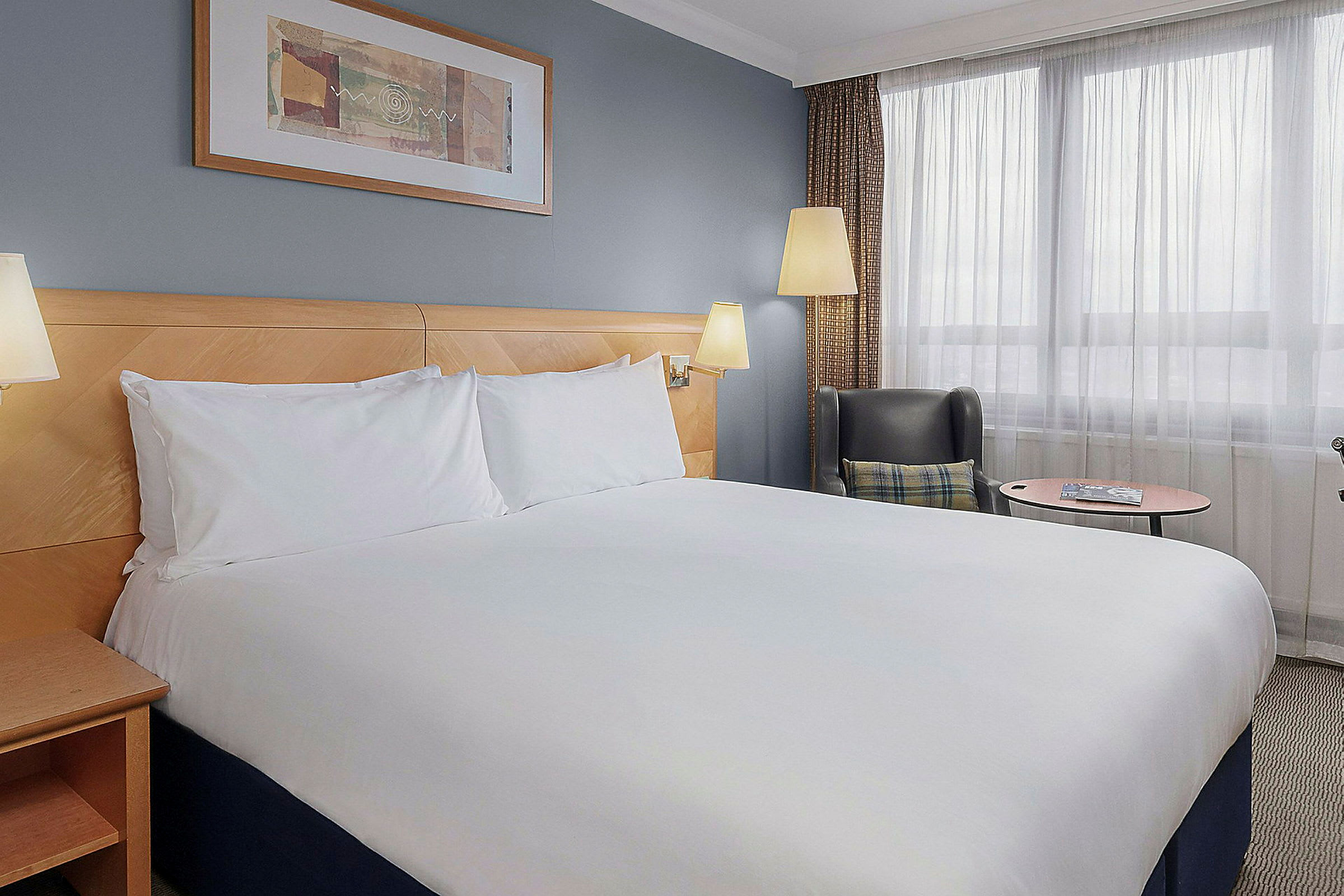 Hotelværelse med dobbeltseng, dekorativ kunst på væggen, en lænestol og et lille bord ved vinduet. (AI-genereret tekst)