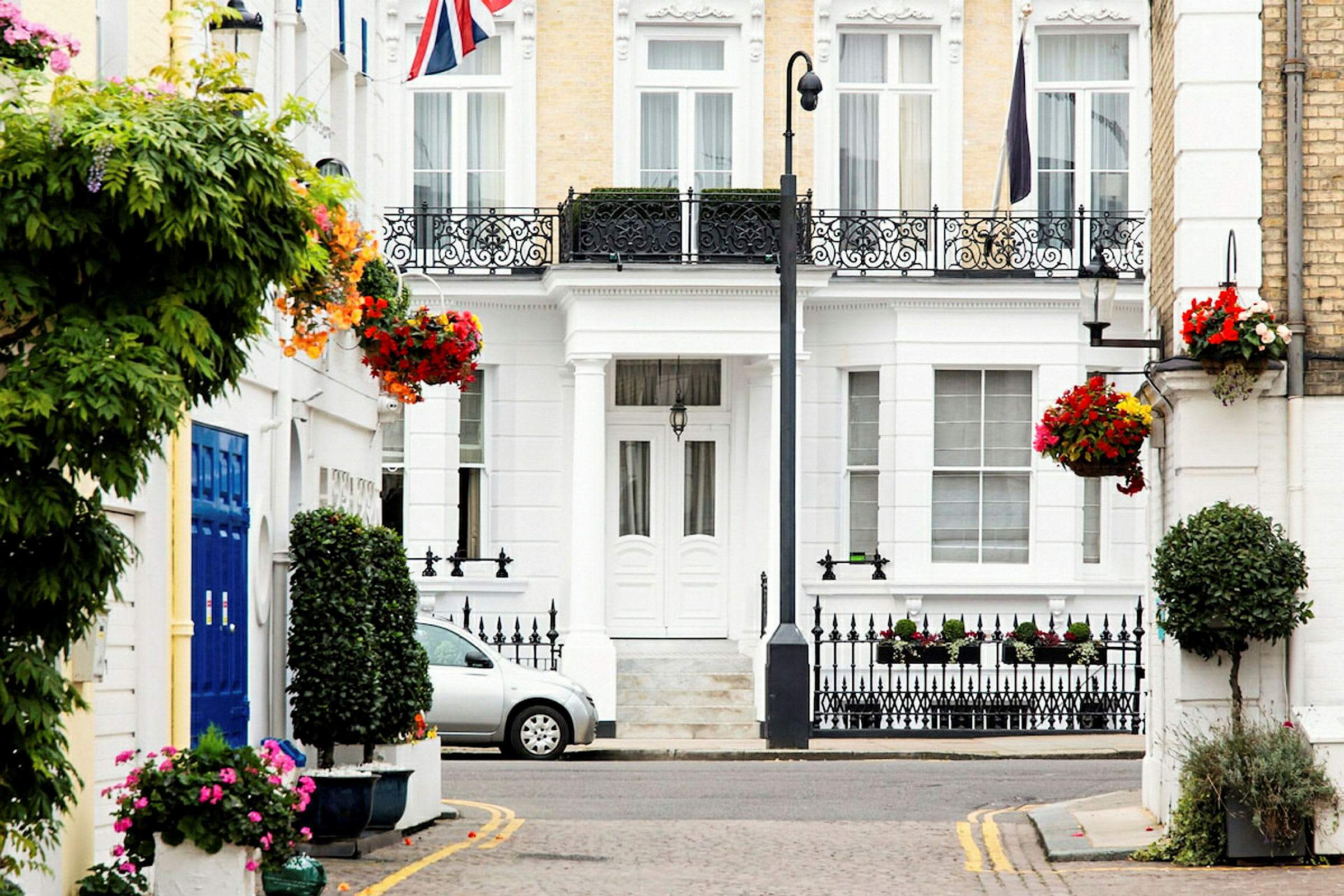 En hvid bygning med dekorative jernrækværk, blomsterdekorationer og en bil parkeret foran. Storbritanniens flag ses. (AI-genereret tekst)