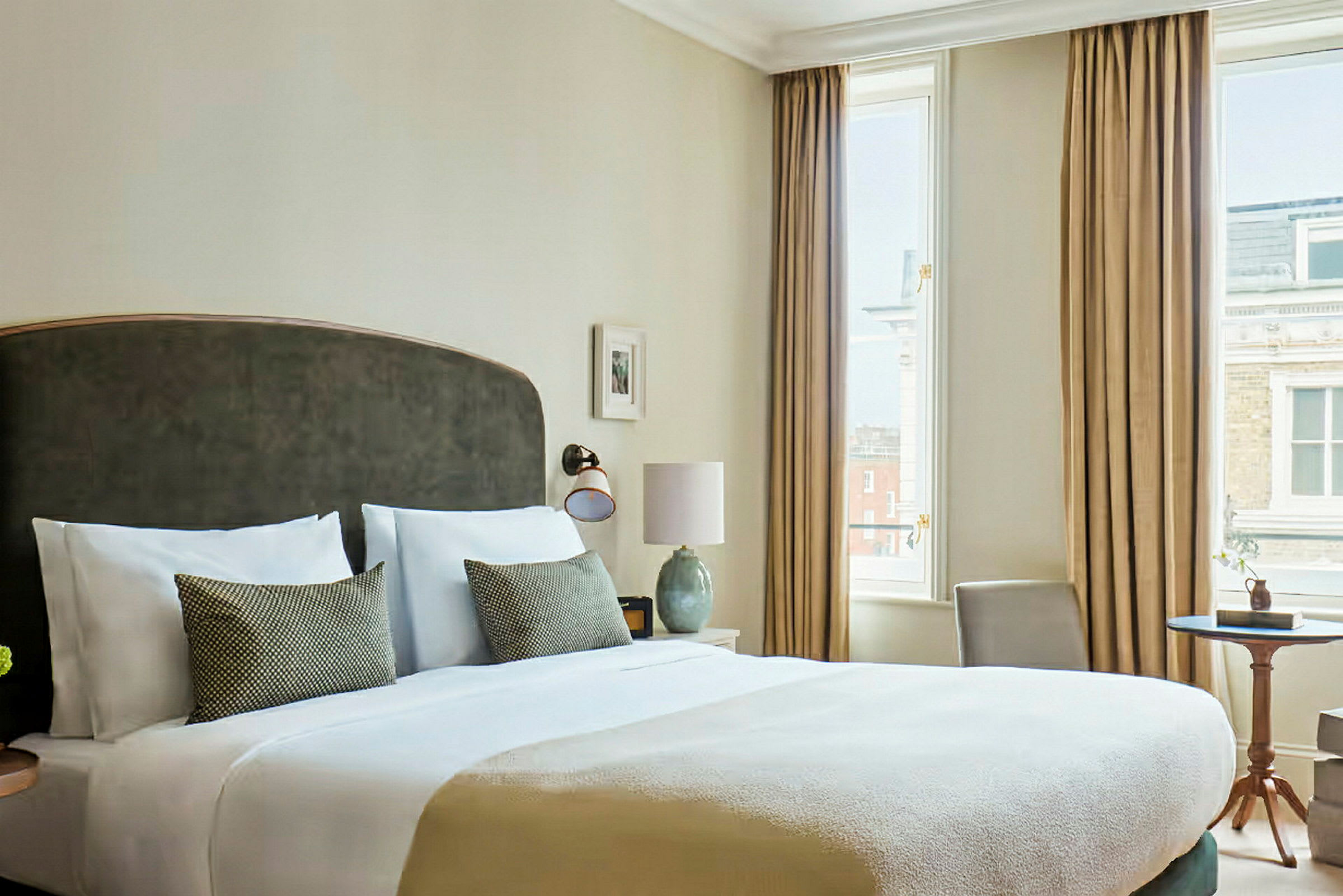 Et elegant hotelværelse med en stor seng, to puder, et natbord og store vinduer med gardiner. (AI-genereret tekst)