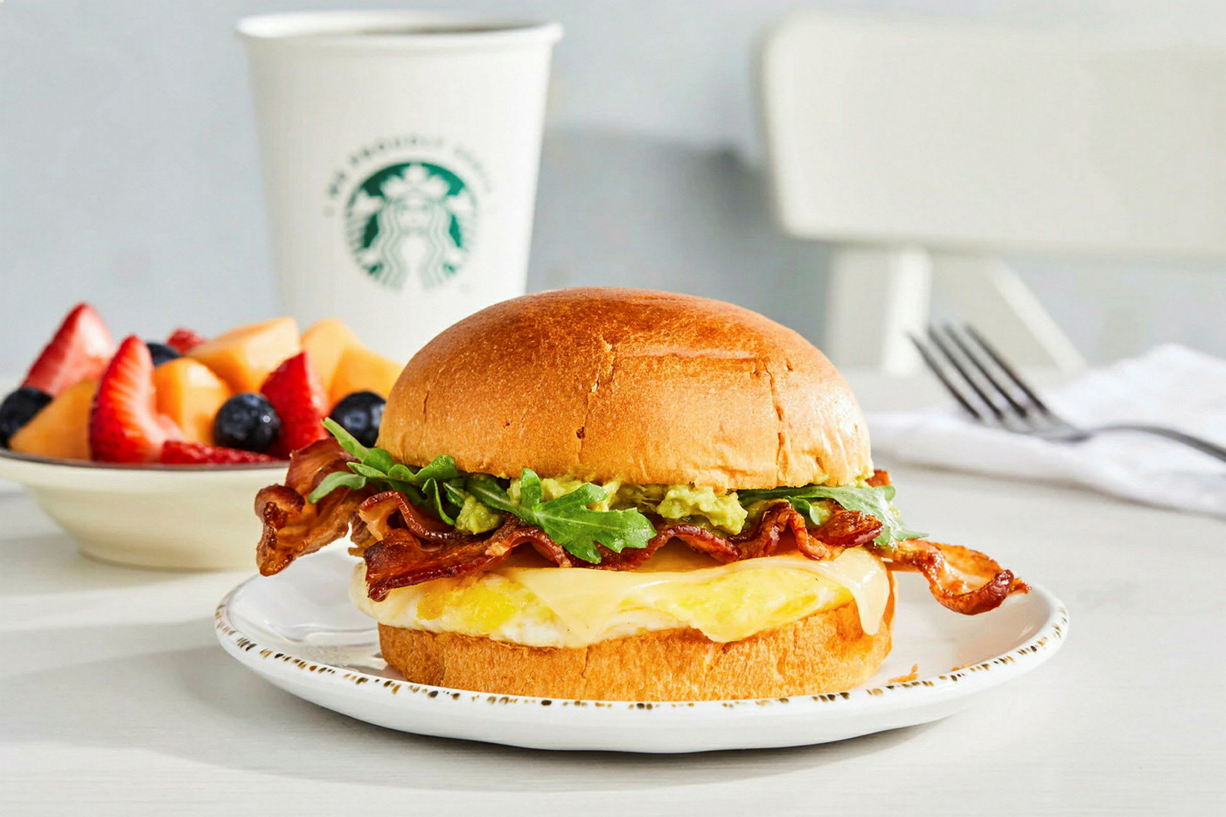 En tallerken med en sandwich, der indeholder bacon, salat, ost og et spejlæg. I baggrunden en kop med logo og en skål med frisk frugt. (AI-genereret tekst)