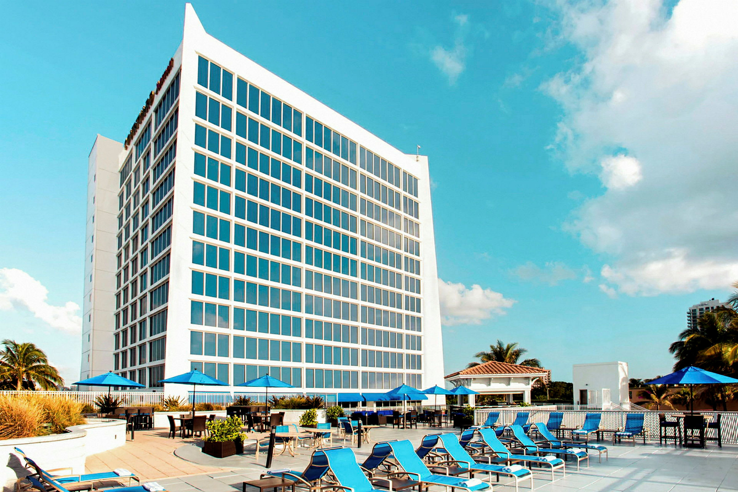 En moderne hotelbygning med store vinduer bagved et poolområde med liggestole og parasoller under en blå himmel. (AI-genereret tekst)