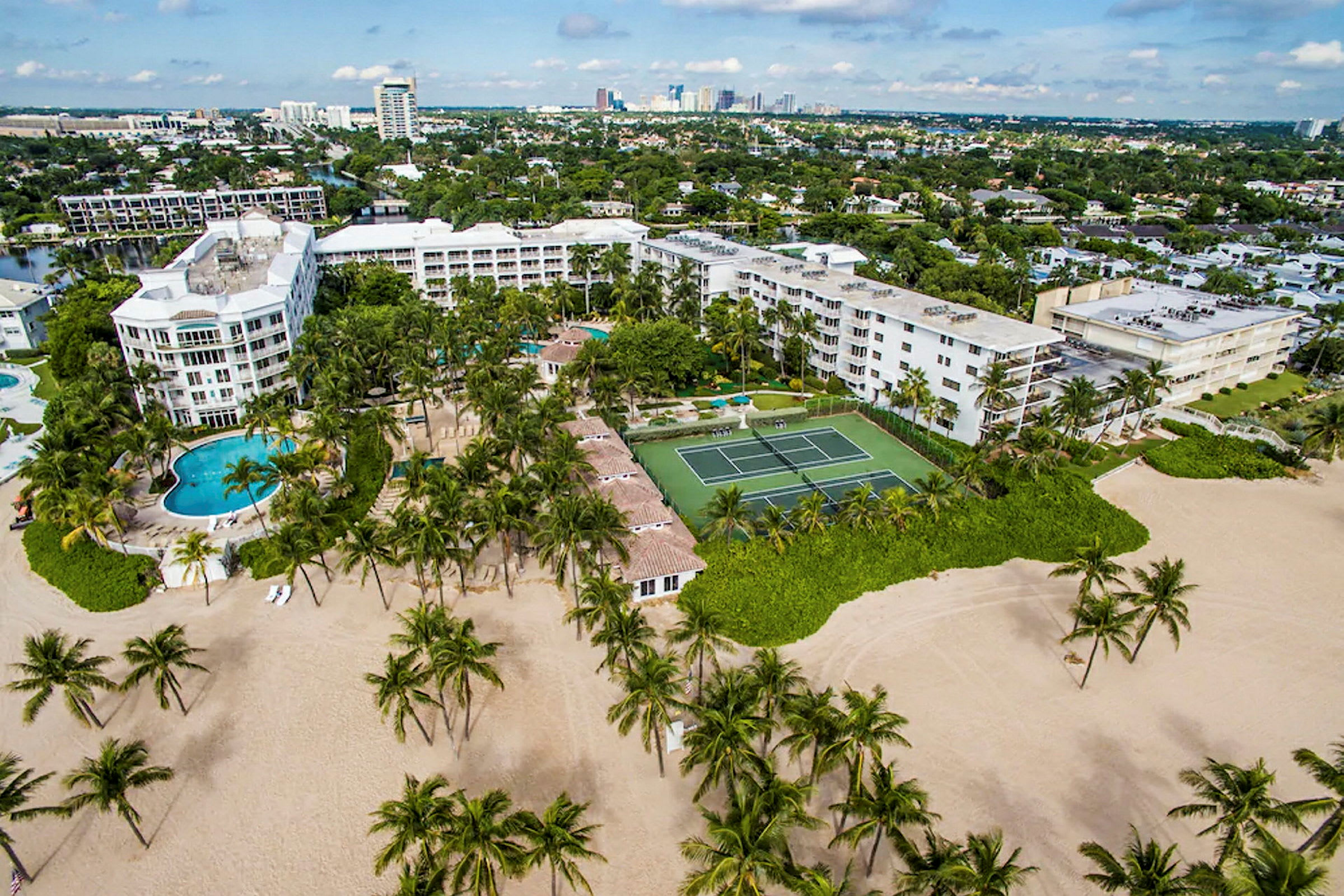 Luftfoto af et feriested med swimmingpools, tennisbane og omkringliggende bygninger beliggende nær en sandstrand med palmer. (AI-genereret tekst)
