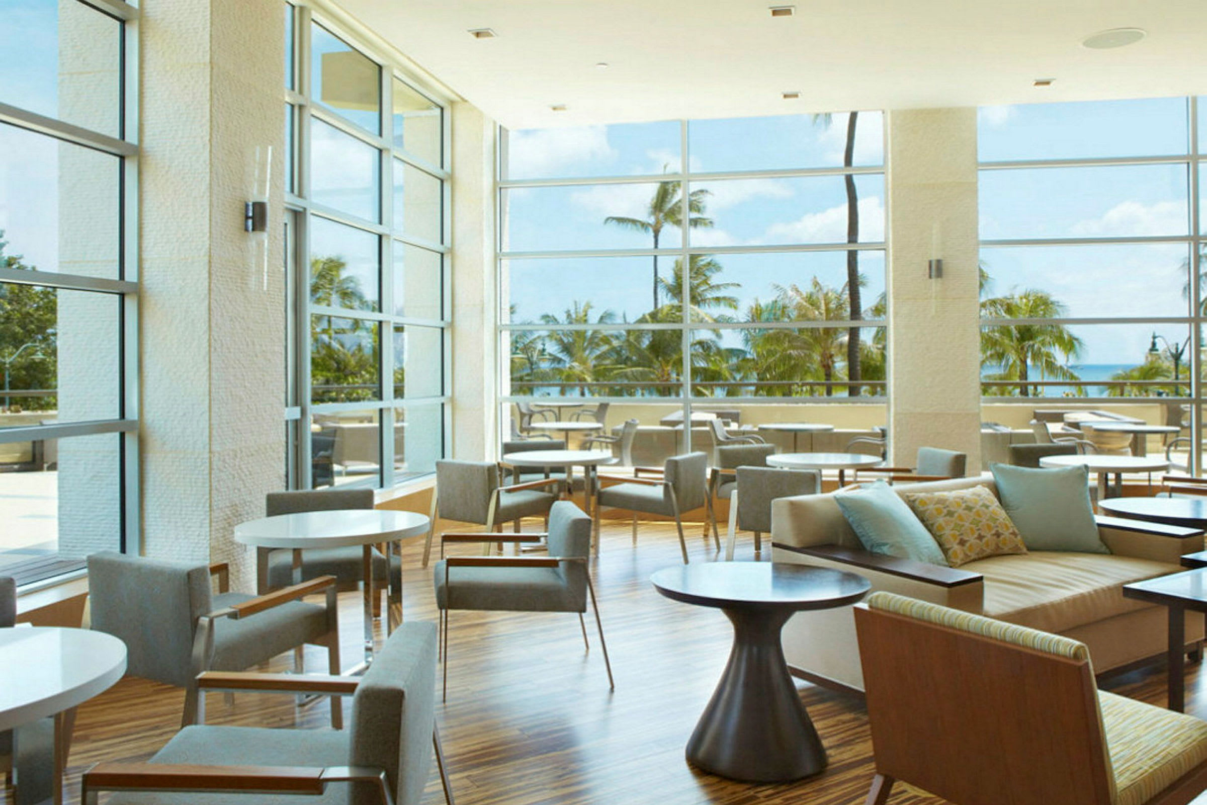 En hotellounge med store glasvinduer, der giver udsigt til palmer og havet. (AI-genereret tekst)