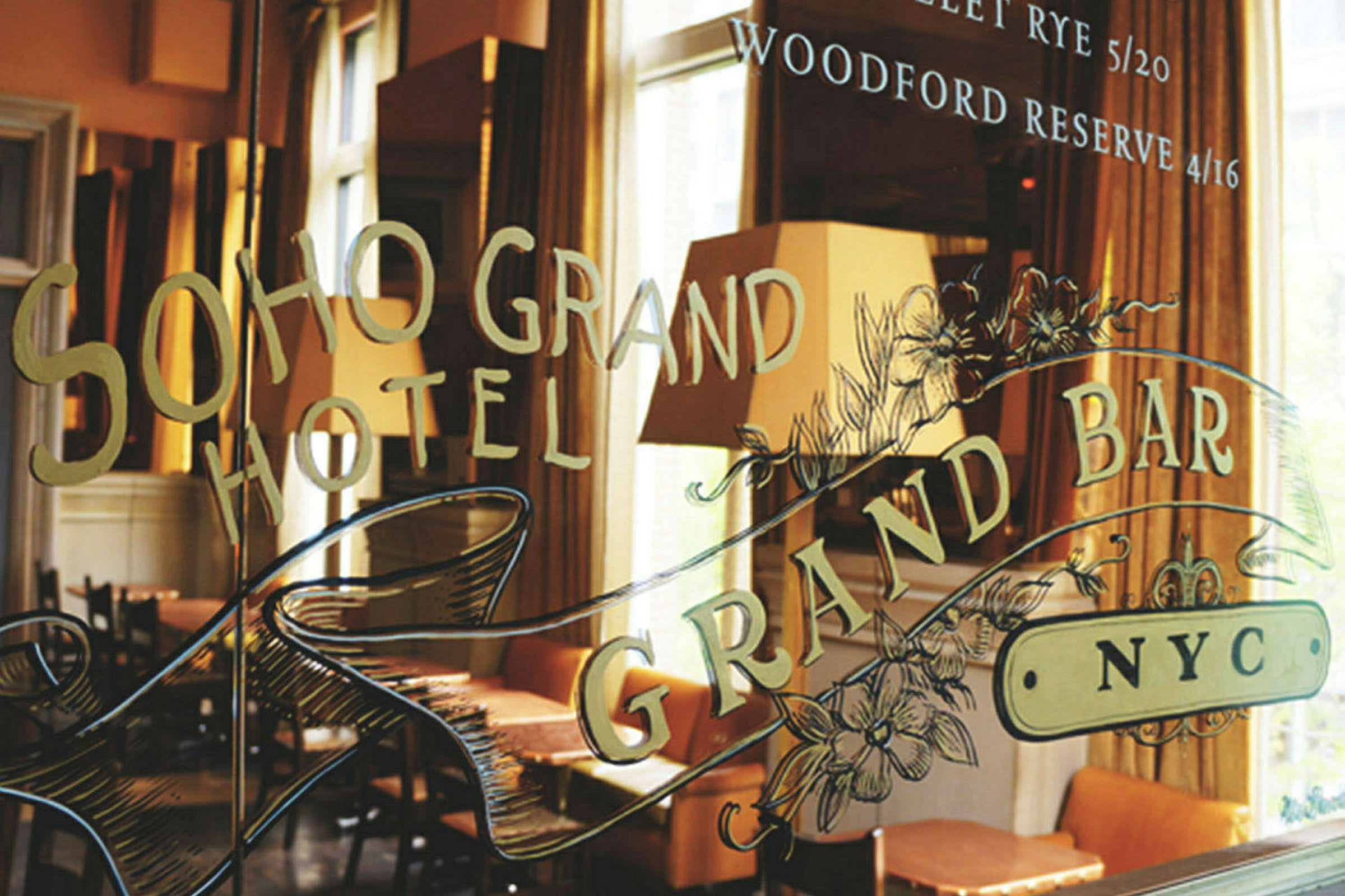 Et vindue med teksten 'Soho Grand Hotel' og 'Grand Bar NYC,' samt dekorationer. Baggrunden viser møbler og lamper. (AI-genereret tekst)