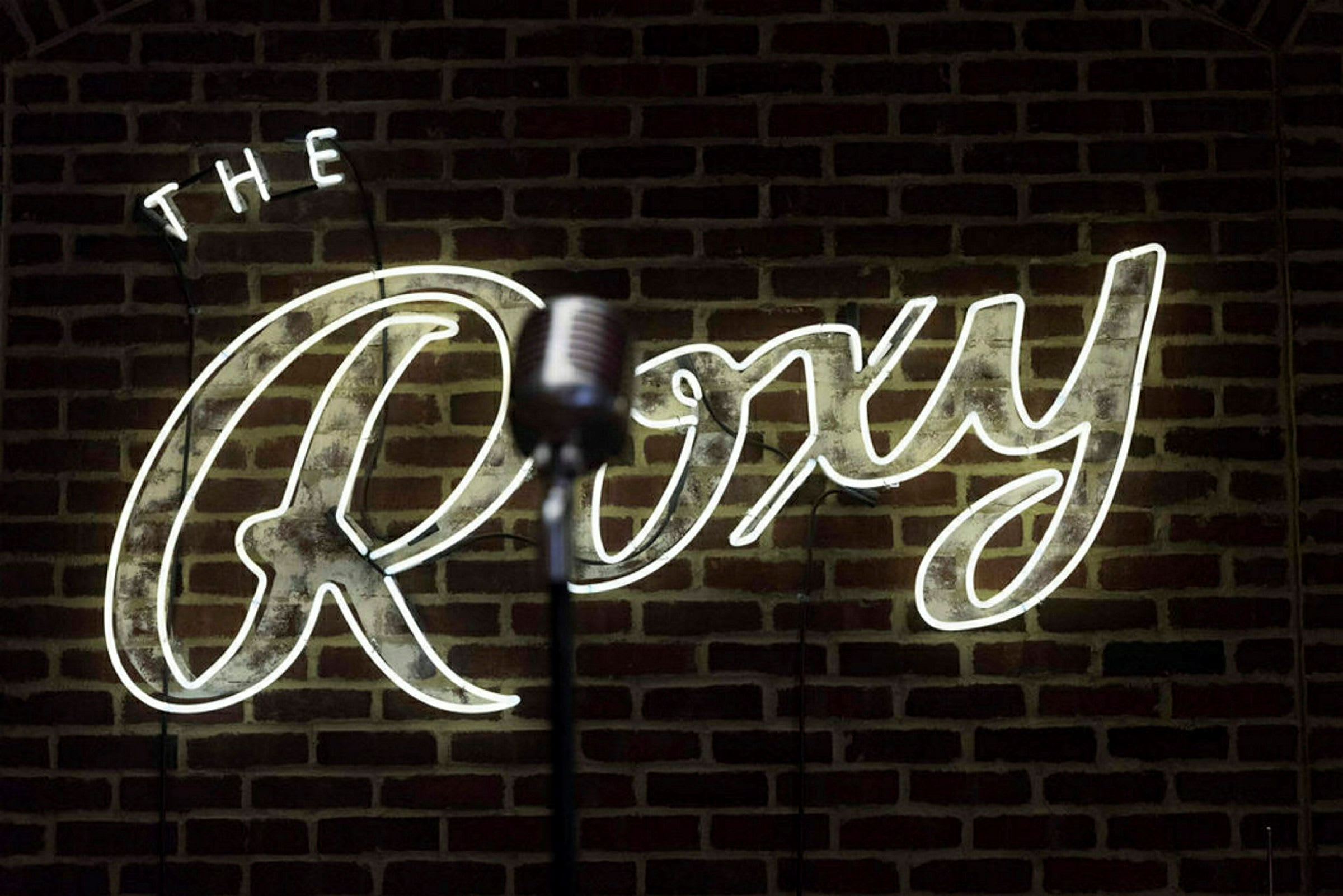 En mikrofon foran et neonskilt med teksten 'The Roxy' på en murstensvæg. (AI-genereret tekst)