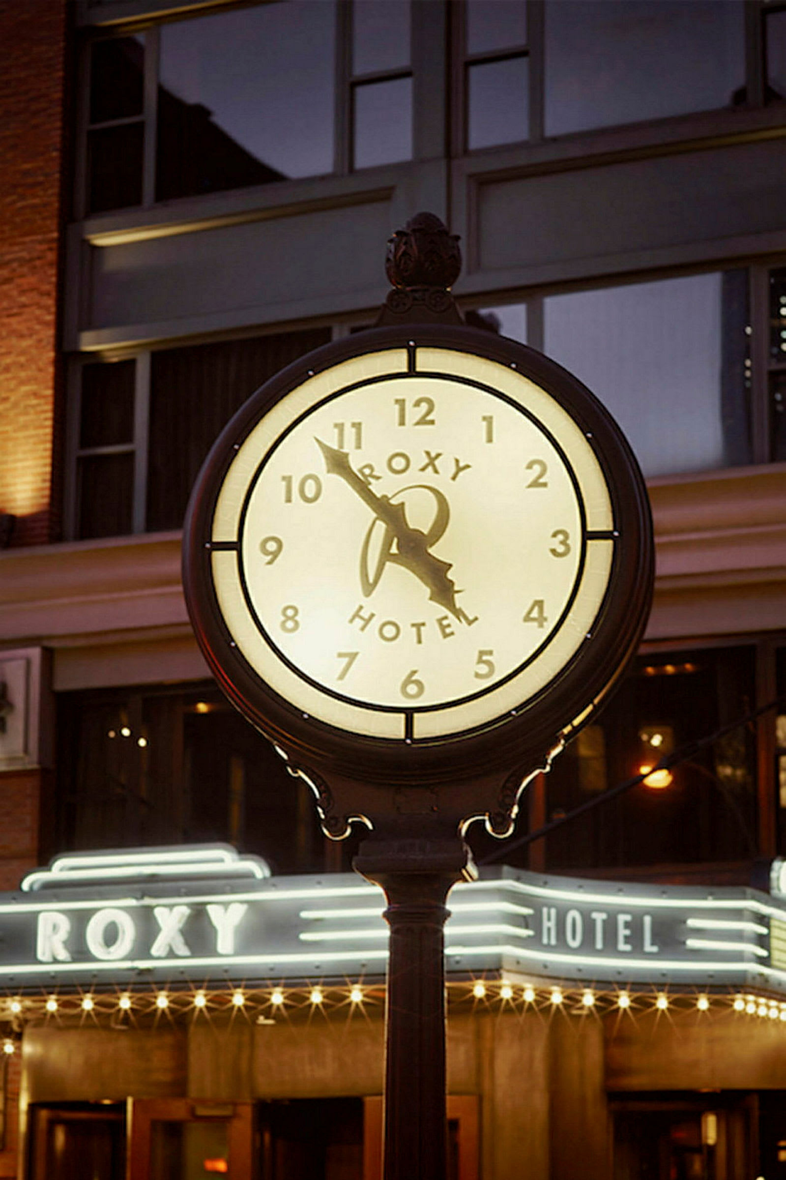 Et oplyst ur med teksten "Roxy Hotel" foran en bygning om aftenen. (AI-genereret tekst)