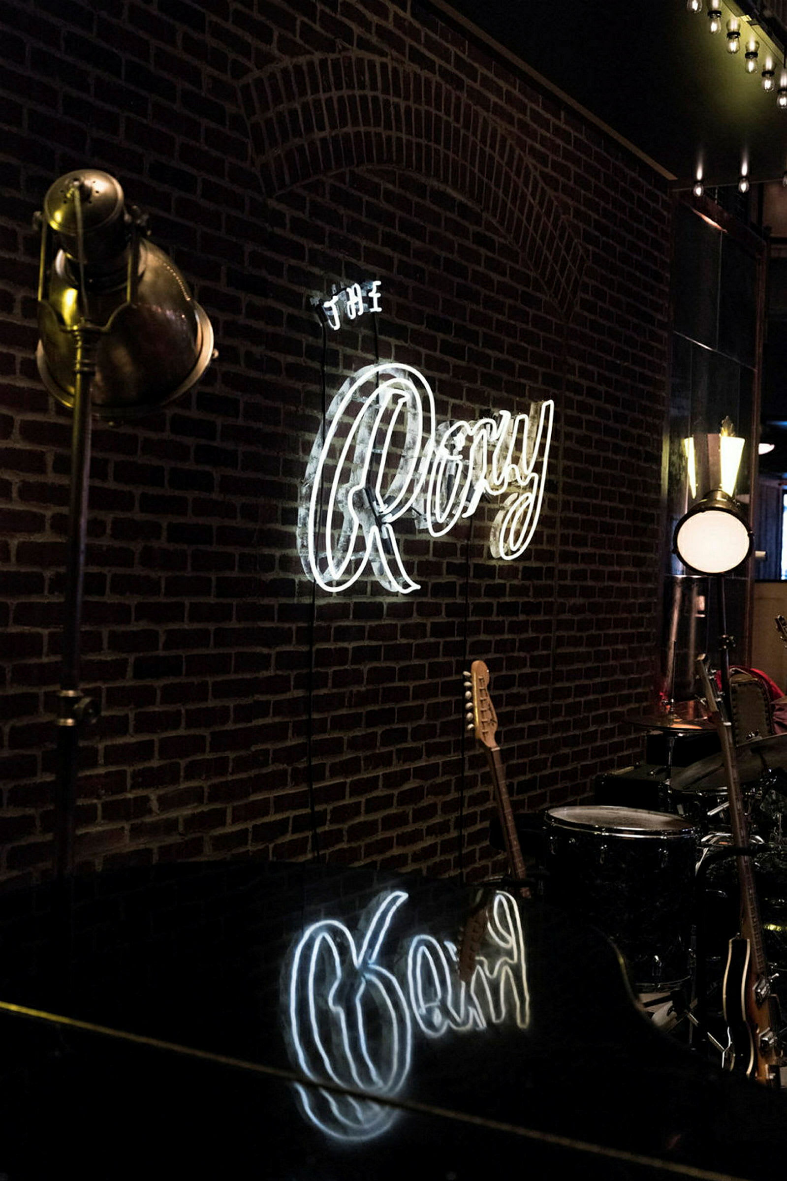 En væg med en neonlampe, der skriver "The Roxy" i hvide bogstaver, samt musikinstrumenter som en guitar og trommer synlige i baggrunden. (AI-genereret tekst)
