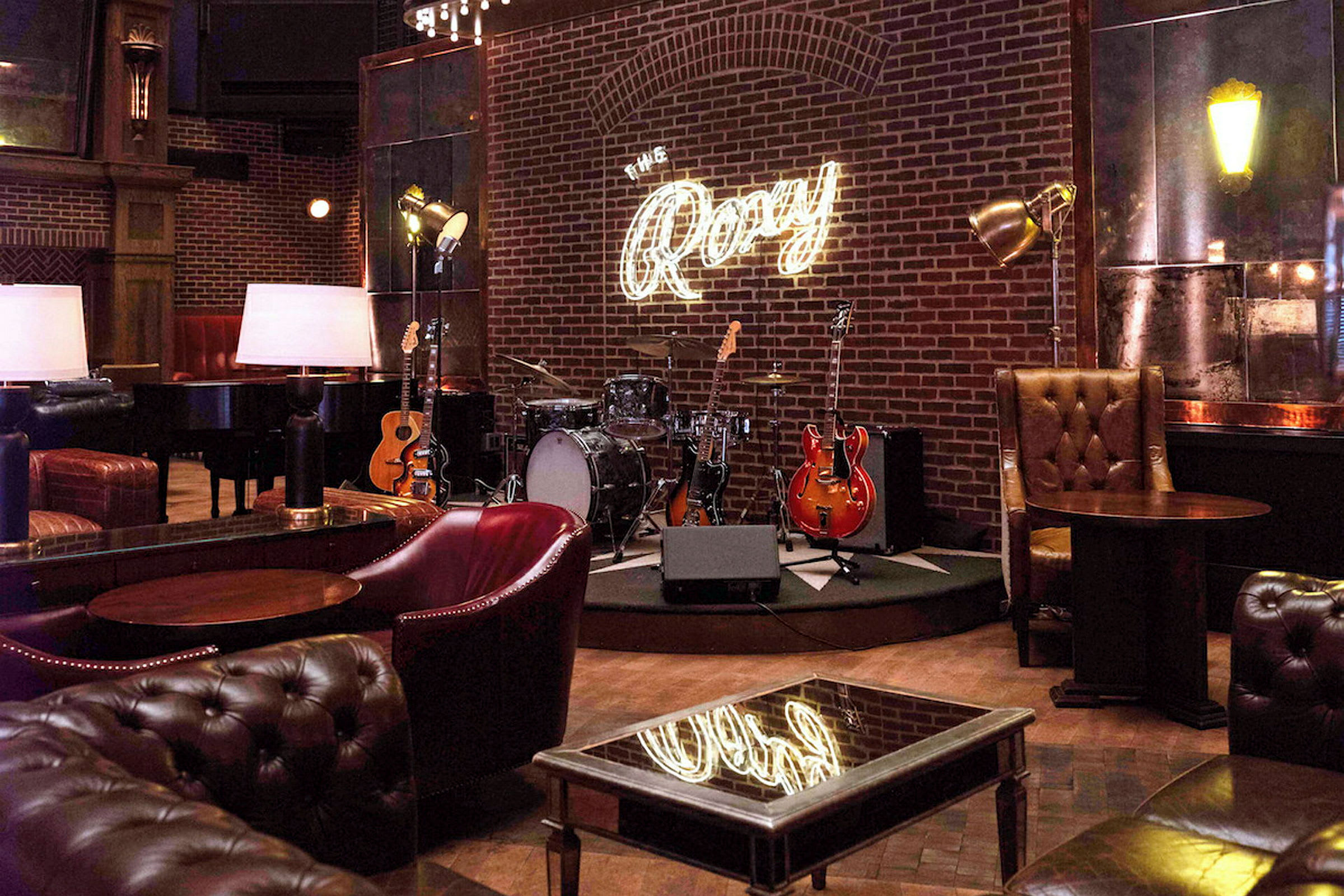 Interiør af en musikklub med guitarer, et trommesæt og en lille scene under et neonskilt, der siger 'The Roxy'. (AI-genereret tekst)