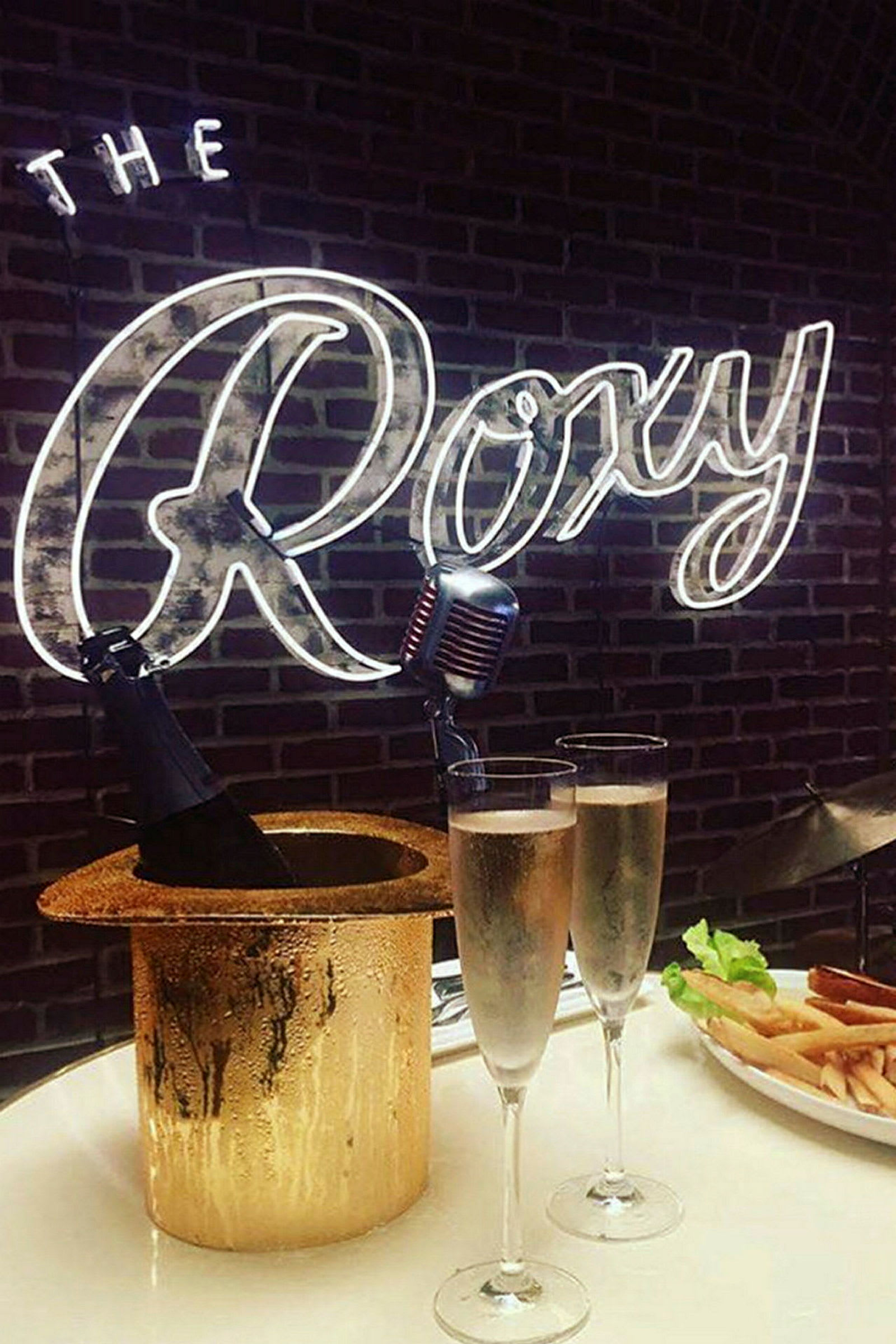 Et bord med to glas mousserende drik, en champagneflaske i en ishink formet som en hat, samt en tallerken med pommes frites. Neonteksten "The Roxy" ses i baggrunden. (AI-genereret tekst)