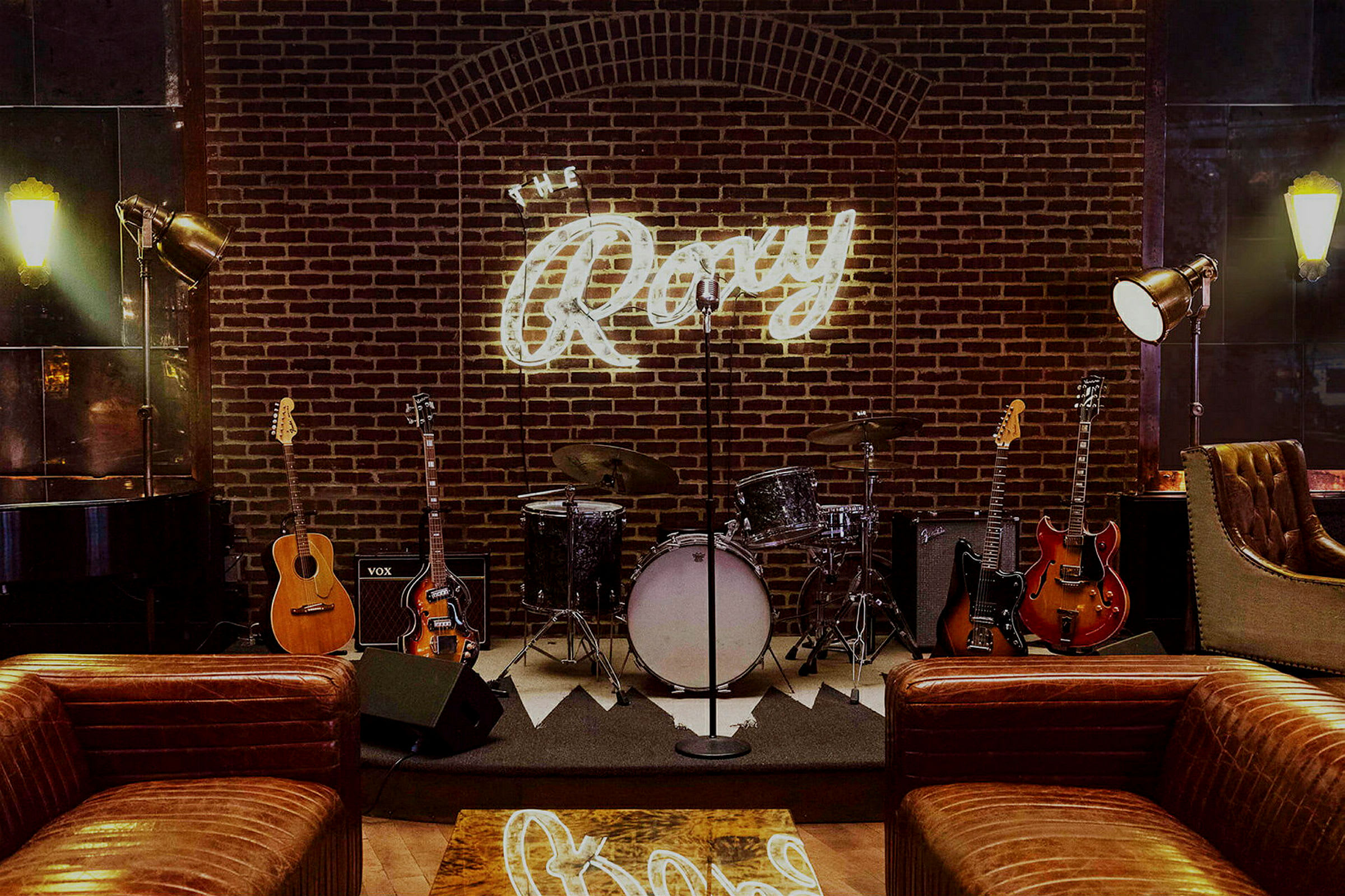En scene med musikinstrumenter som trommer og guitarer foran en murstensvæg med et lysende skilt, der siger "The Roxy". (AI-genereret tekst)