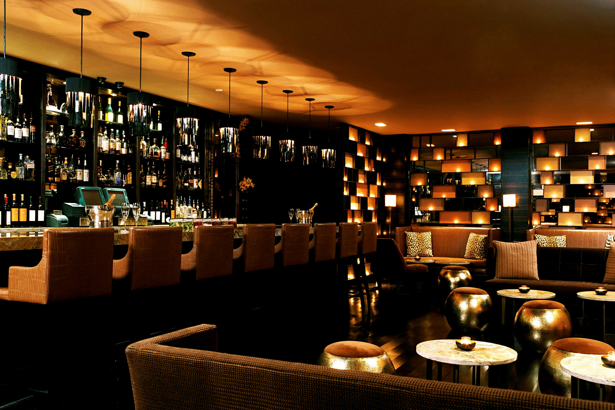 En elegant bar med højryggede stole ved bardisken, flasker i baggrunden samt sofaer og borde med blød belysning. (AI-genereret tekst)